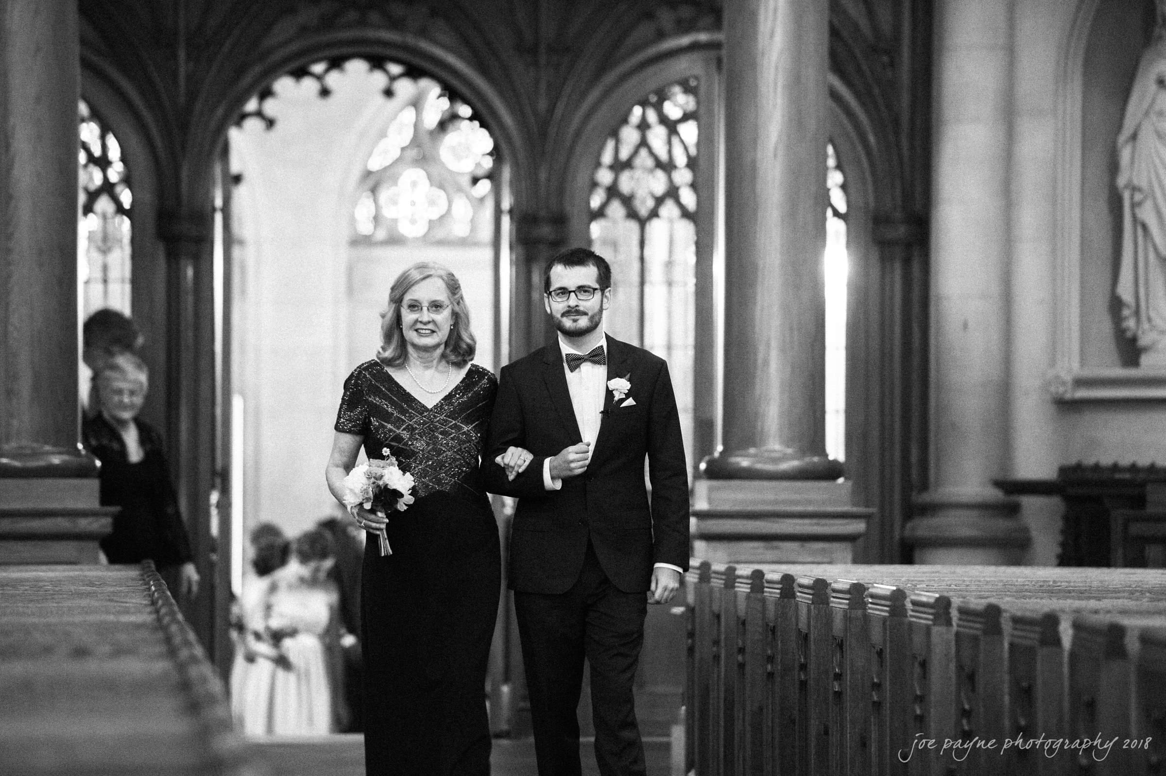 A woman and a man, dressed formally, walk arm in arm down the aisle of a gothic-style church. The woman holds a bouquet. The background features ornate columns and stained-glass windows.