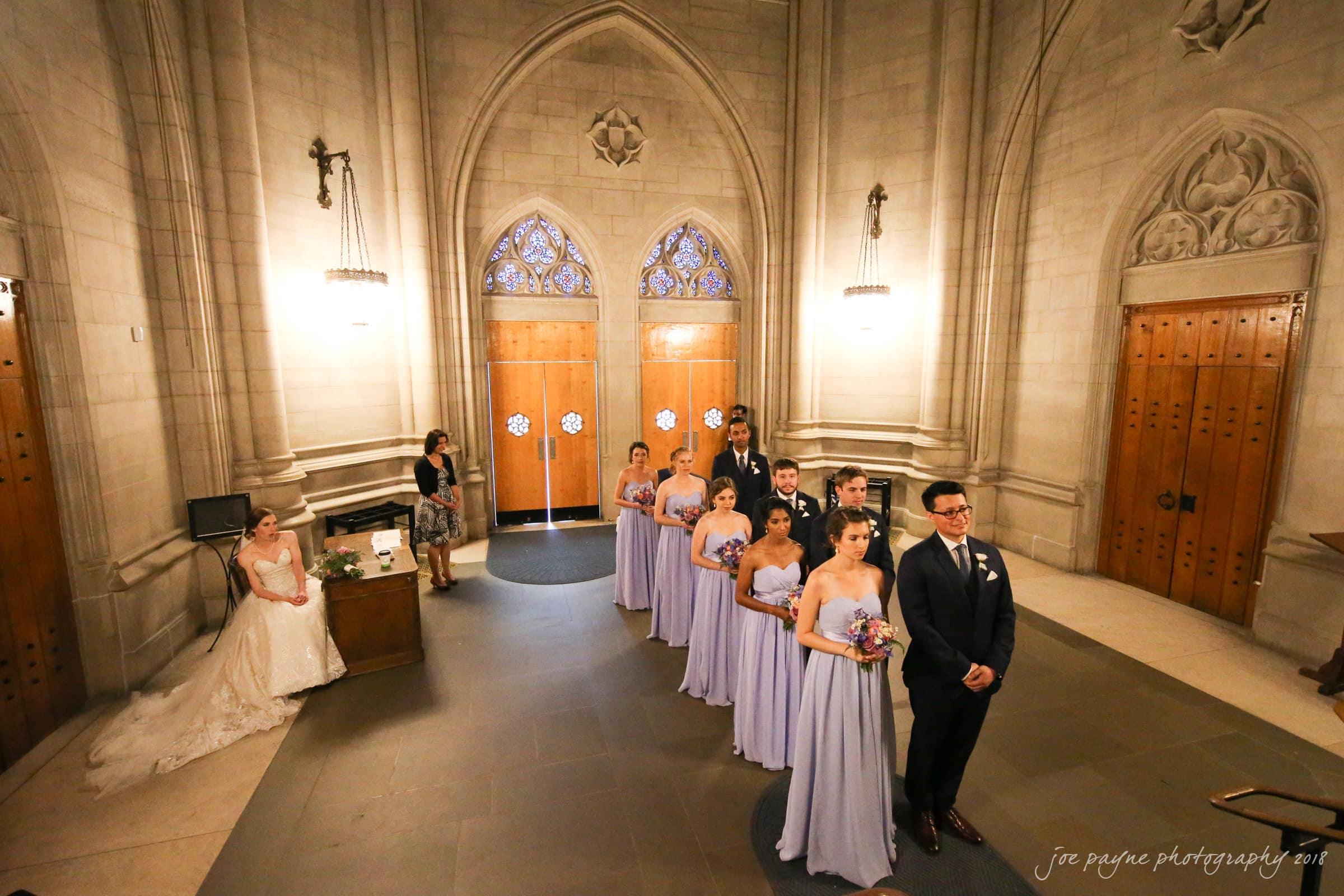A wedding party stands in a cathedral-like setting. The bridesmaids in lavender gowns and the groomsmen in dark suits line up in pairs. The bride, in a white gown, is seated nearby. The setting is grand, with arched doors and ornate details.