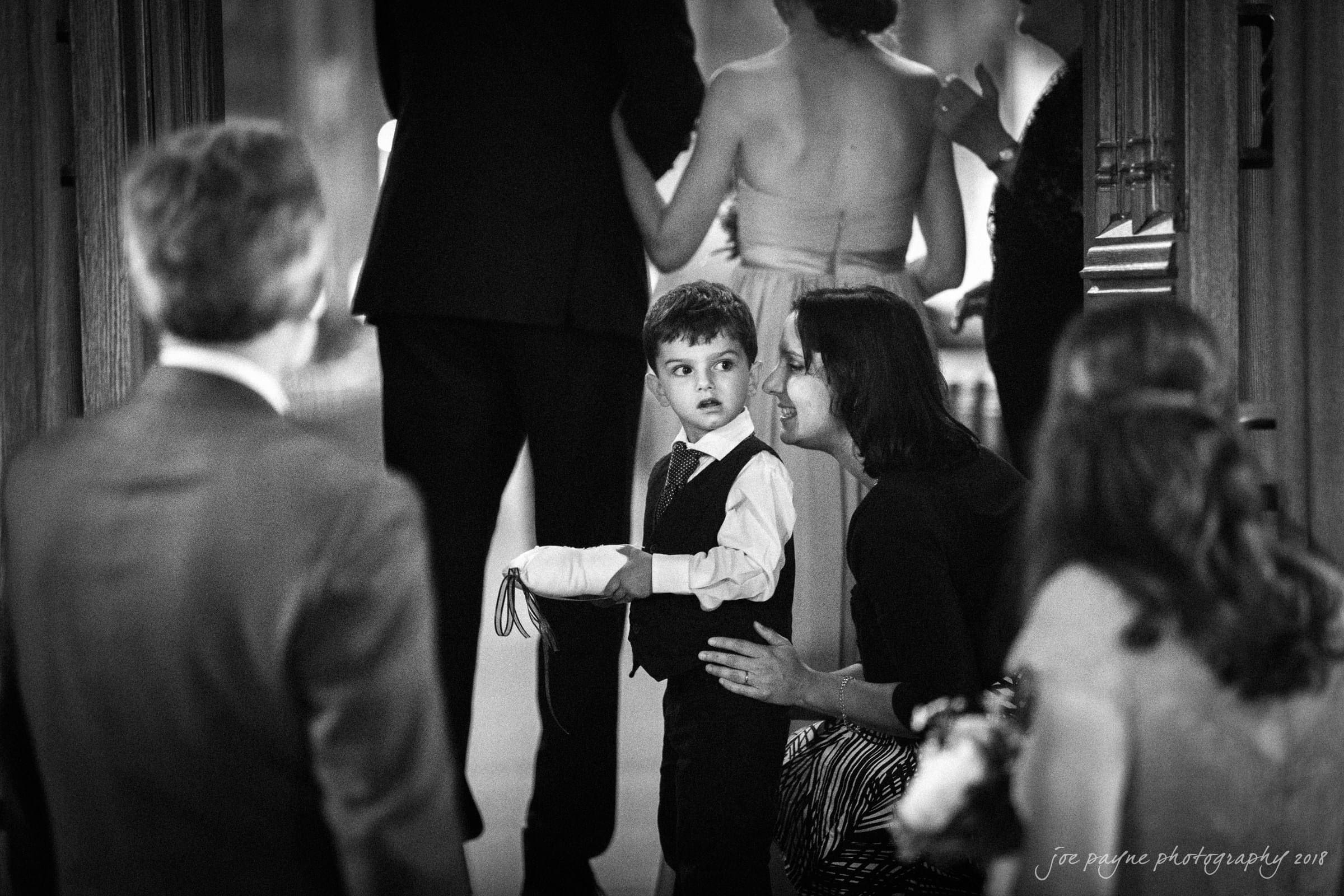 A young boy in formal attire holds rings on a pillow. A woman crouches beside him, smiling, as other adults stand in the background, dressed for an event. The setting appears to be indoors, possibly a wedding. The image is in black and white.