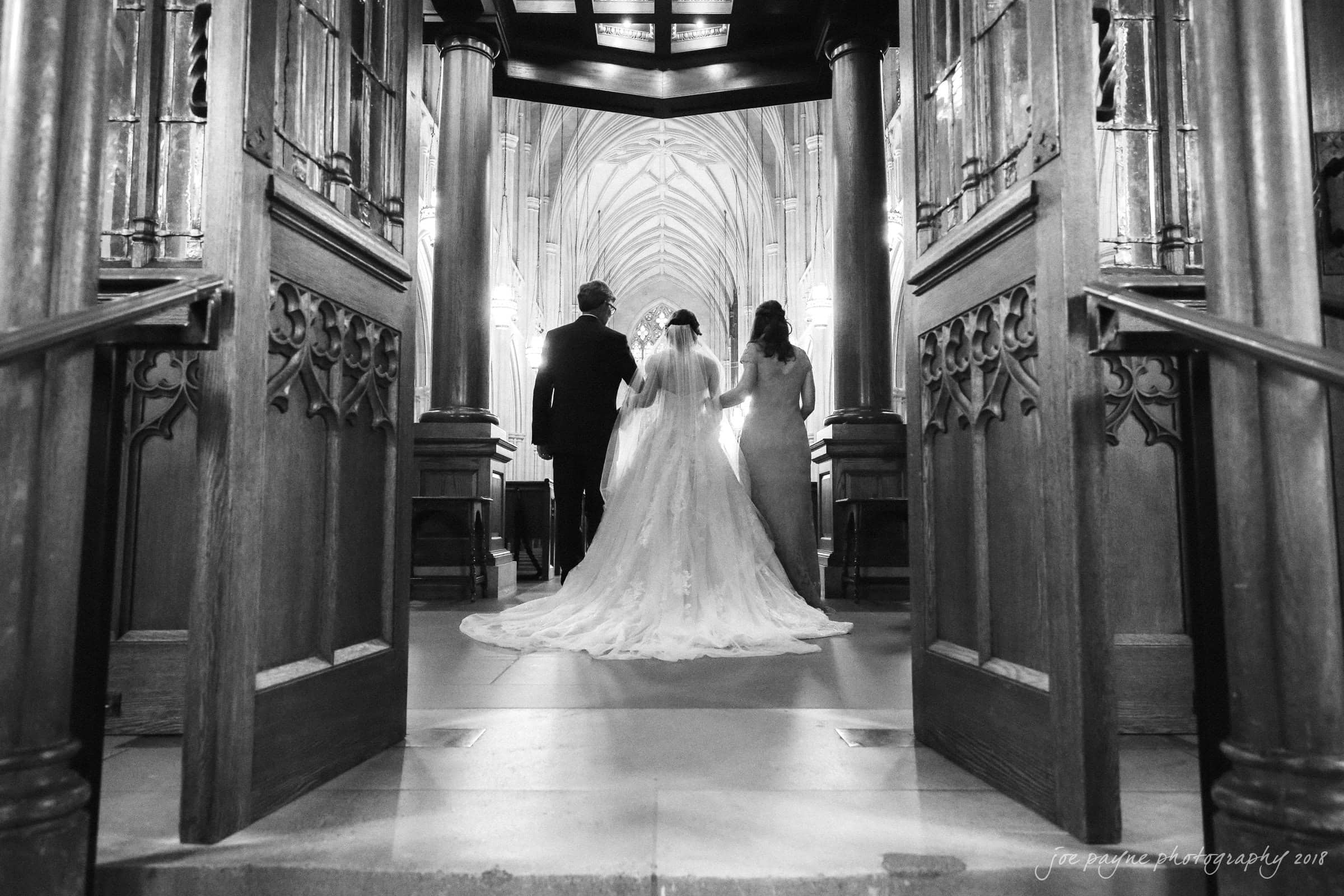 A black and white photo shows three people walking down the aisle of a grand cathedral. They are dressed in formal attire, with one person on each side of a bride. The open wooden doors frame the intricately designed interior ahead.