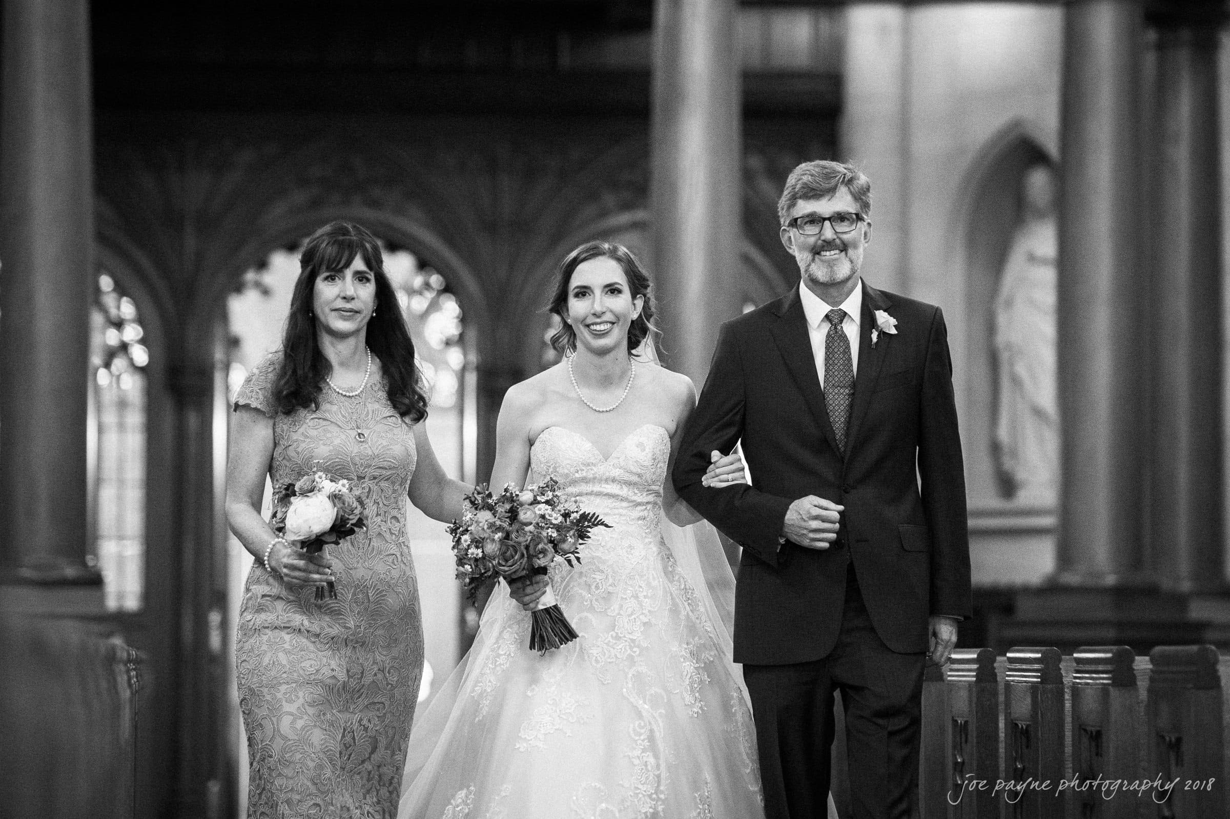 A bride in a strapless gown holding a bouquet is walking down the aisle between two people in formal attire, a woman in a lace dress and a man in a suit. They are in a church setting with ornate arches in the background.