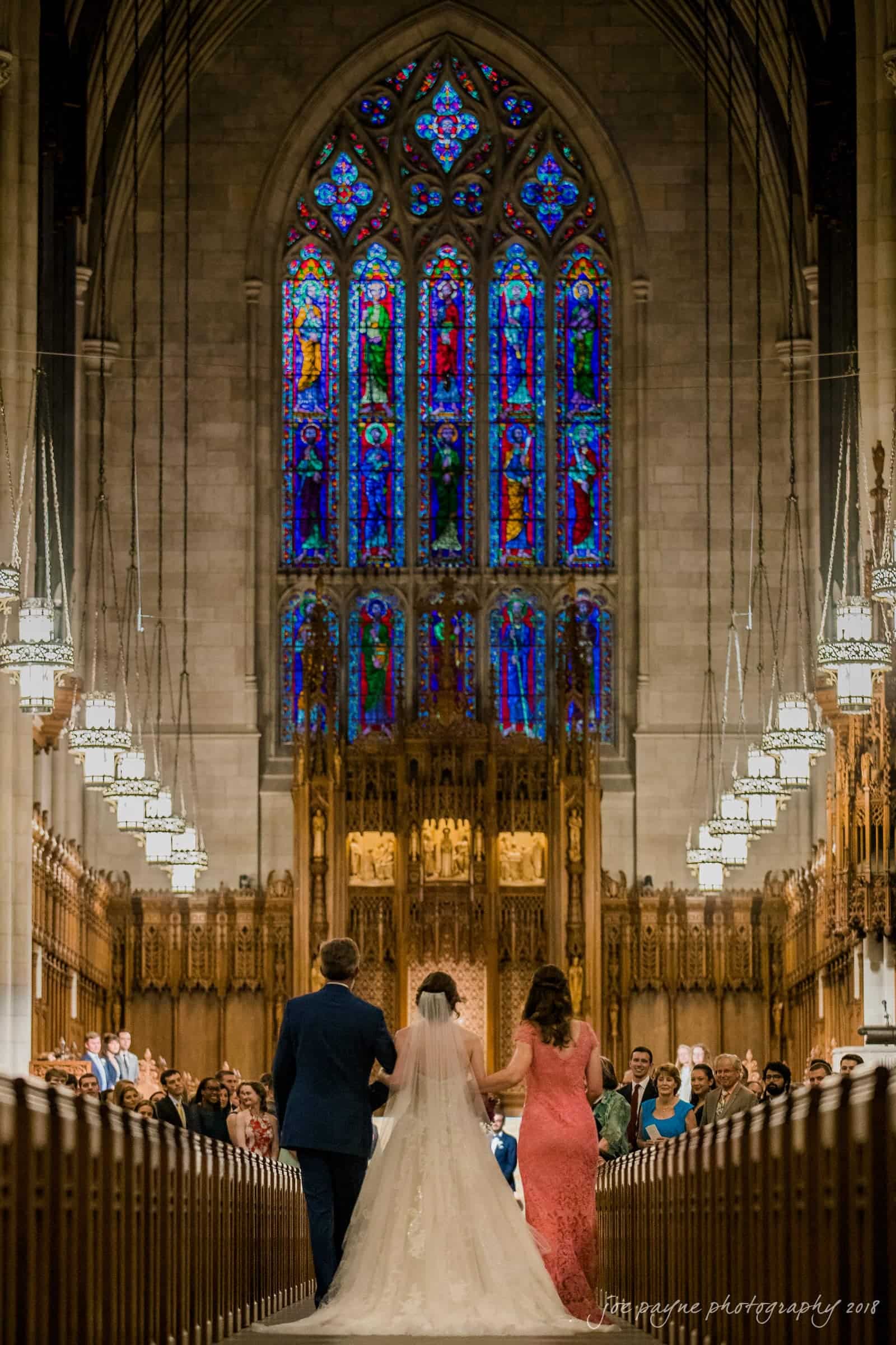 A bride walks down the aisle between two people in a grand cathedral with intricate stained glass windows and ornate woodwork. The guests are seated on either side, and the venue is decorated with hanging lights.