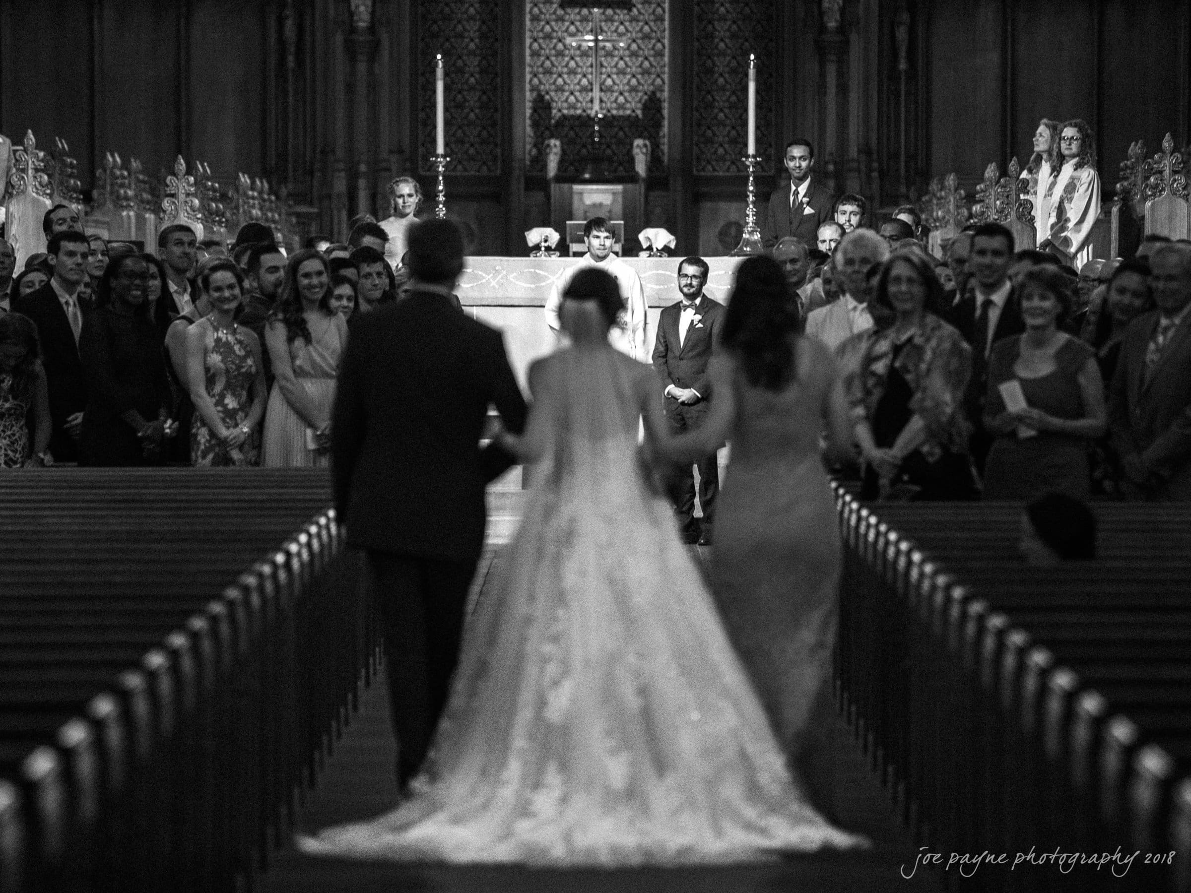 A bride with her parents walks down the aisle of a grand cathedral filled with guests. The groom waits at the altar, flanked by attendants, against the backdrop of ornate architectural details.