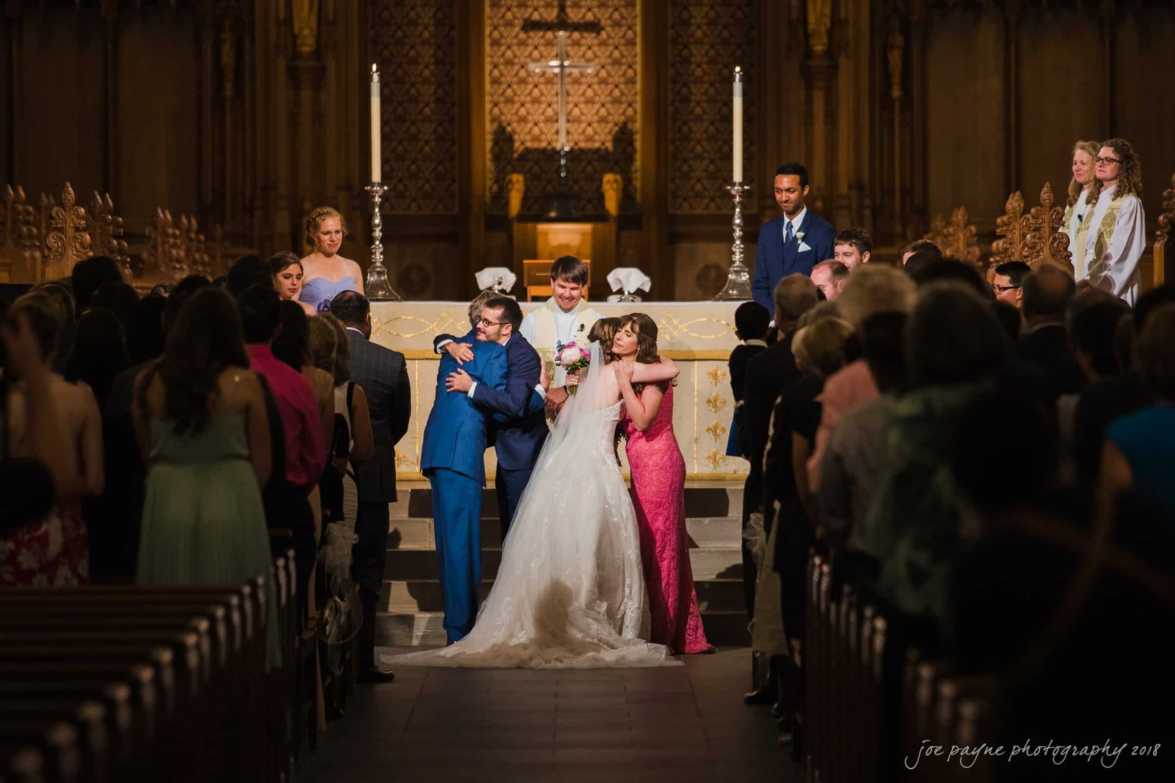 A wedding ceremony in a church. The bride and groom stand at the altar, embracing family members. The wedding party and guests are gathered, watching the scene. The setting is warmly lit, with ornate wooden details in the background.