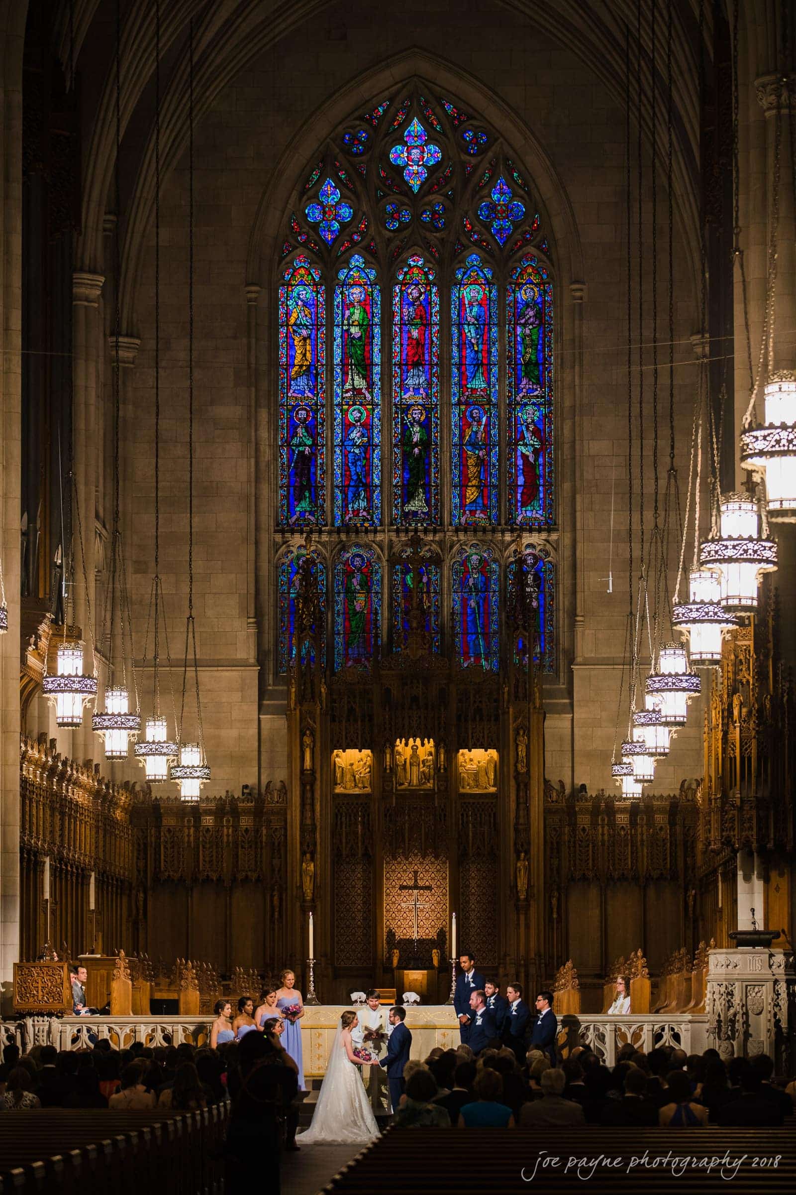 A couple stands at the altar in a grand cathedral with high ceilings and ornate stained-glass windows. Wedding guests are seated, and the space is illuminated by hanging chandeliers.