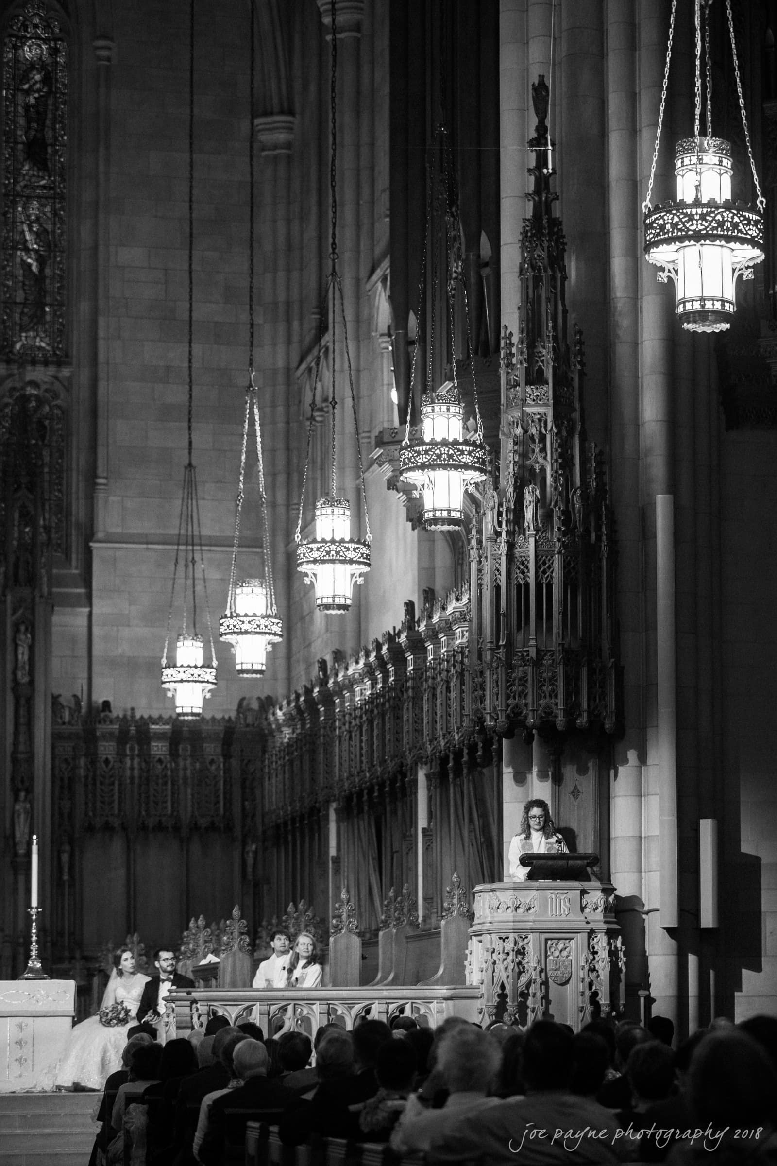 A black-and-white photo of a cathedral interior during a ceremony. A speaker at the pulpit addresses a seated congregation. Ornate chandeliers hang from the ceiling, and intricate woodwork decorates the walls.
