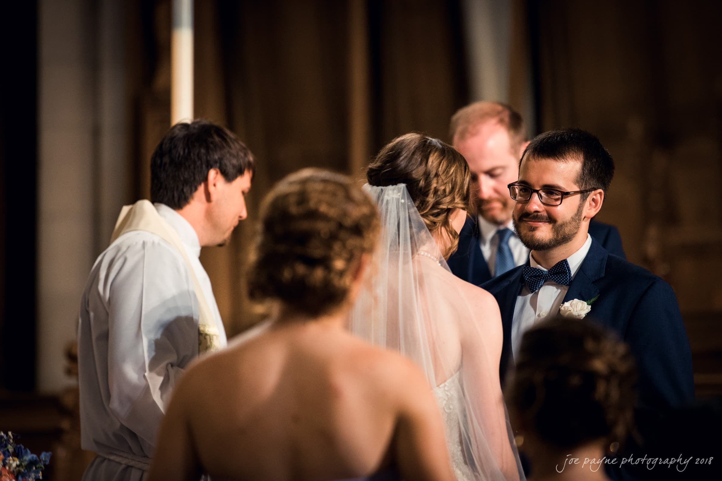 A groom in a blue suit and bow tie smiles at the bride during a wedding ceremony. The bride, wearing a veil, stands with her back to the camera, while a priest in white officiates. Bridesmaids, slightly out of focus, are in the foreground.