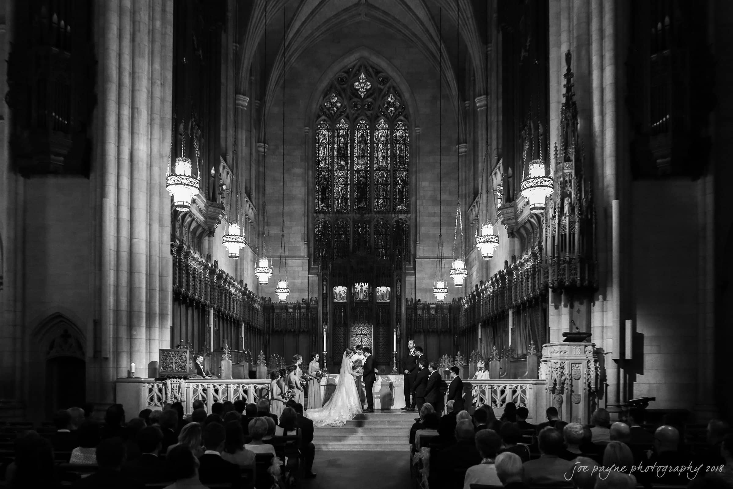 A black and white image of a wedding ceremony inside a grand cathedral. The bride and groom stand at the altar, framed by ornate architecture and tall columns. Guests are seated in pews, witnessing the event.