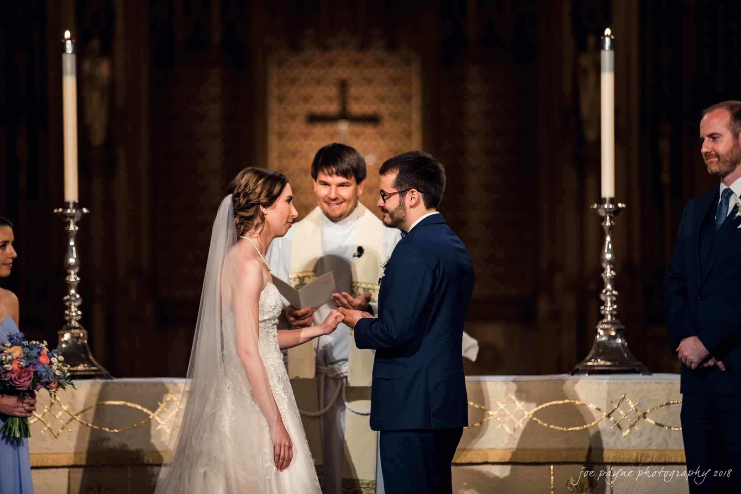 A bride and groom exchange rings during a wedding ceremony at an altar, as a smiling officiant stands behind them. The bride wears a white gown, and the groom is in a blue suit. Two attendants stand nearby, one holding a bouquet.