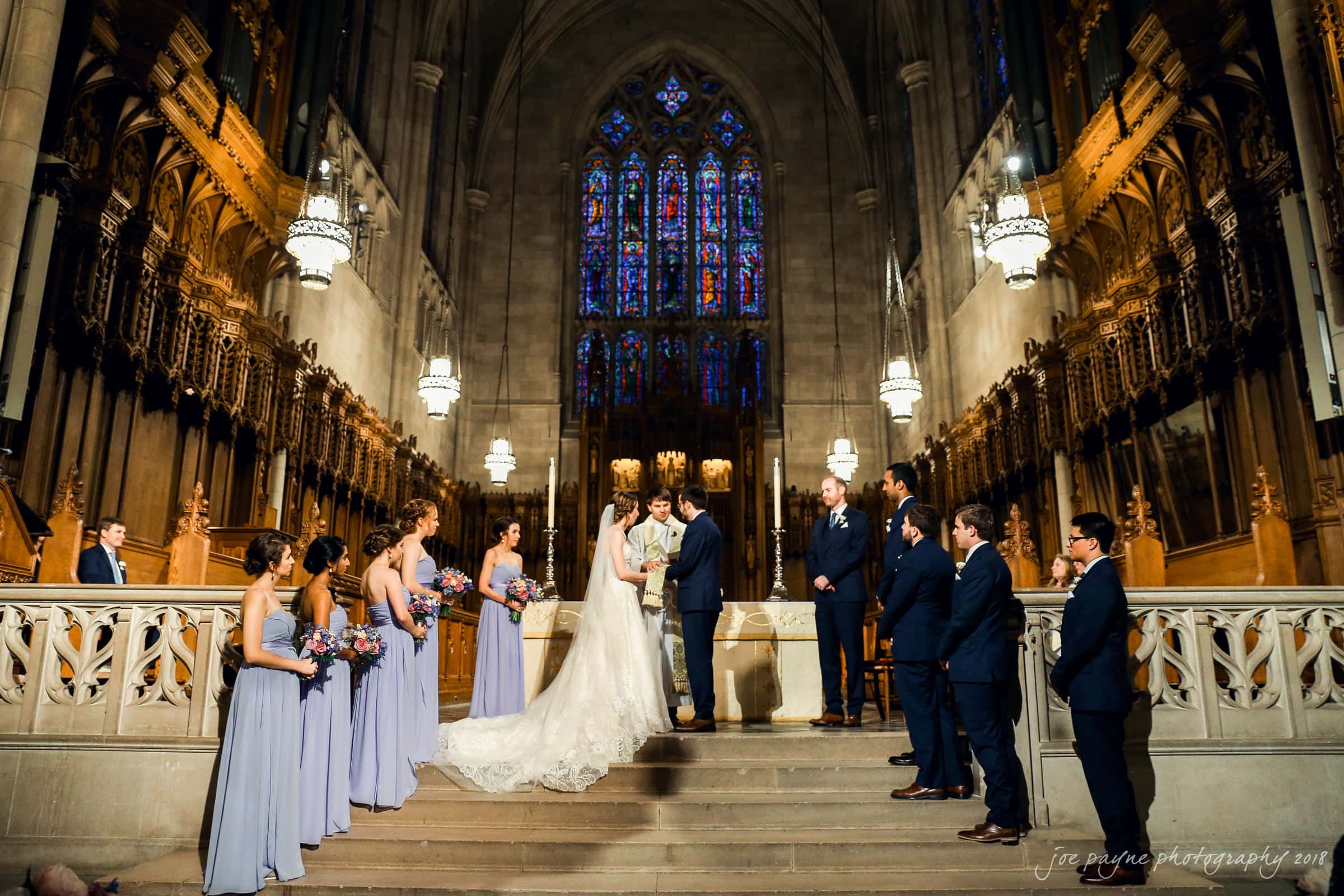 A wedding ceremony inside a grand cathedral, with a couple at the altar. Bridesmaids in lavender dresses and groomsmen in dark suits stand on either side. Stained glass windows and ornate architecture are visible in the background.