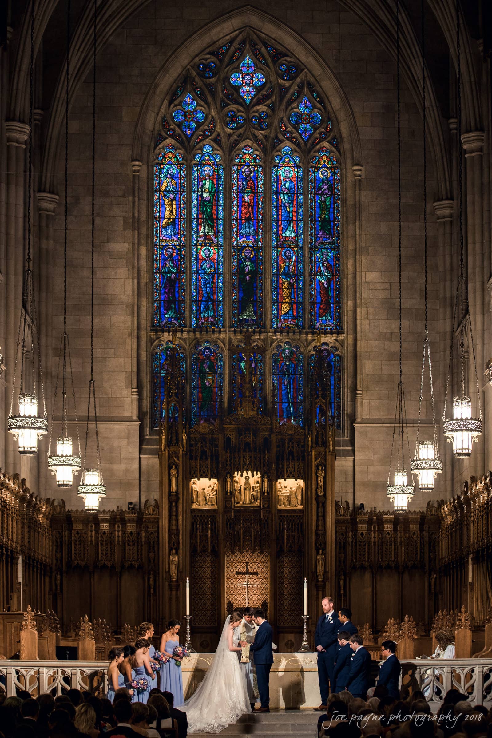 A wedding ceremony taking place in a grand cathedral with a large, colorful stained glass window. The bride and groom stand at the altar, surrounded by the bridal party, with ornate lighting and architecture in the background.