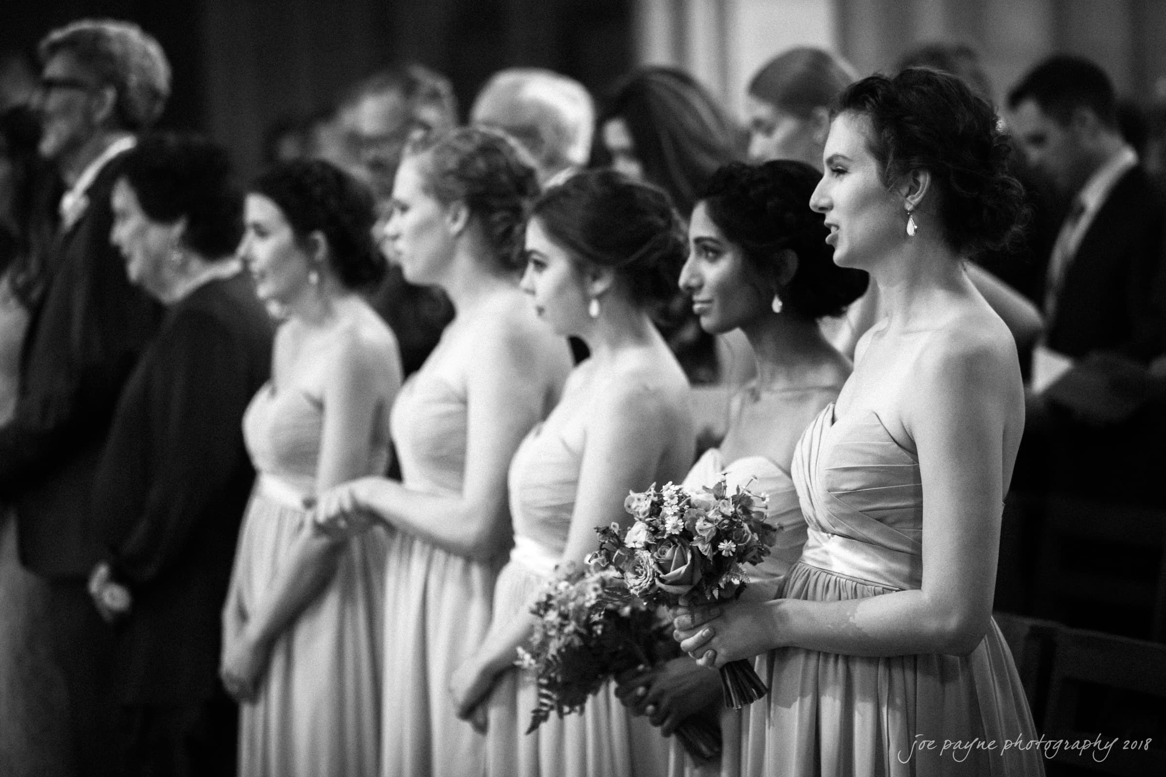 A group of bridesmaids in strapless dresses stand in a row during a ceremony. One holds a bouquet of flowers. The background is blurred, showing other seated guests. The photo is in black and white.