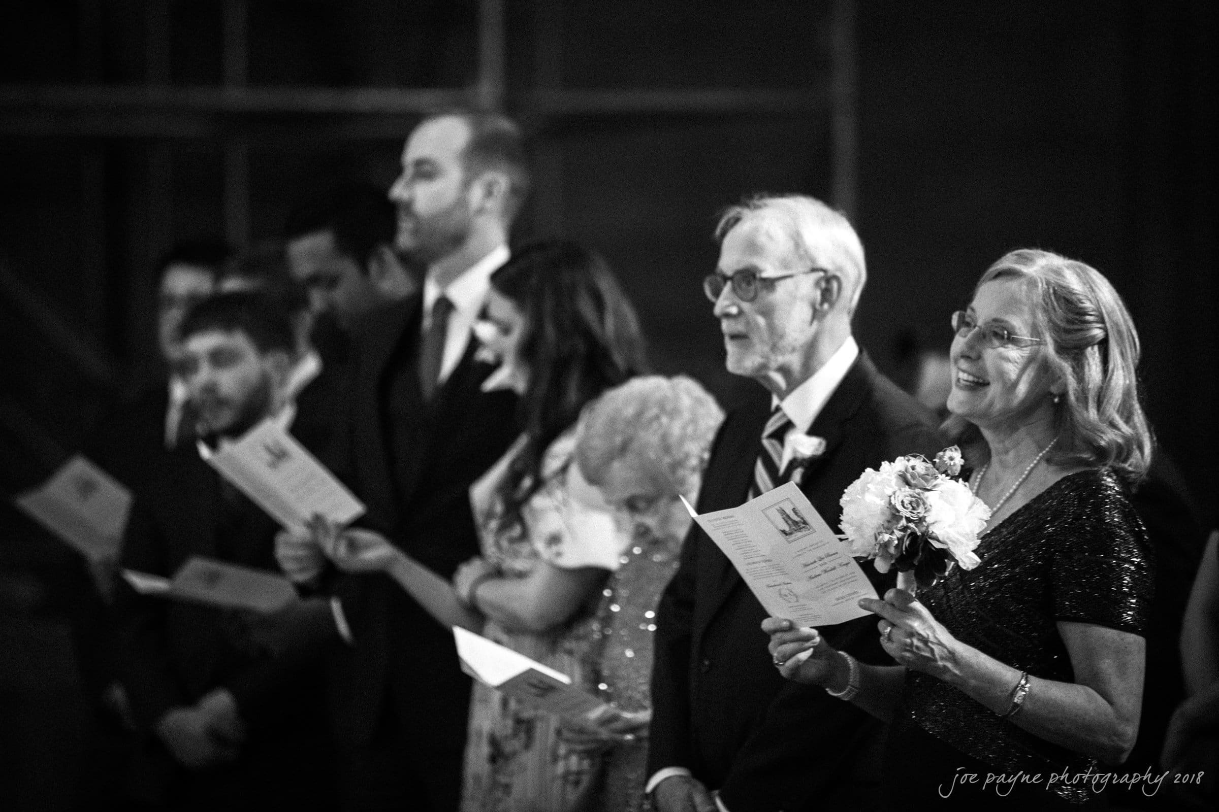 A group of people standing in a row, dressed formally. Some hold papers, smiling, in what appears to be a ceremony or event setting. The scene is in black and white, showing a joyful atmosphere.