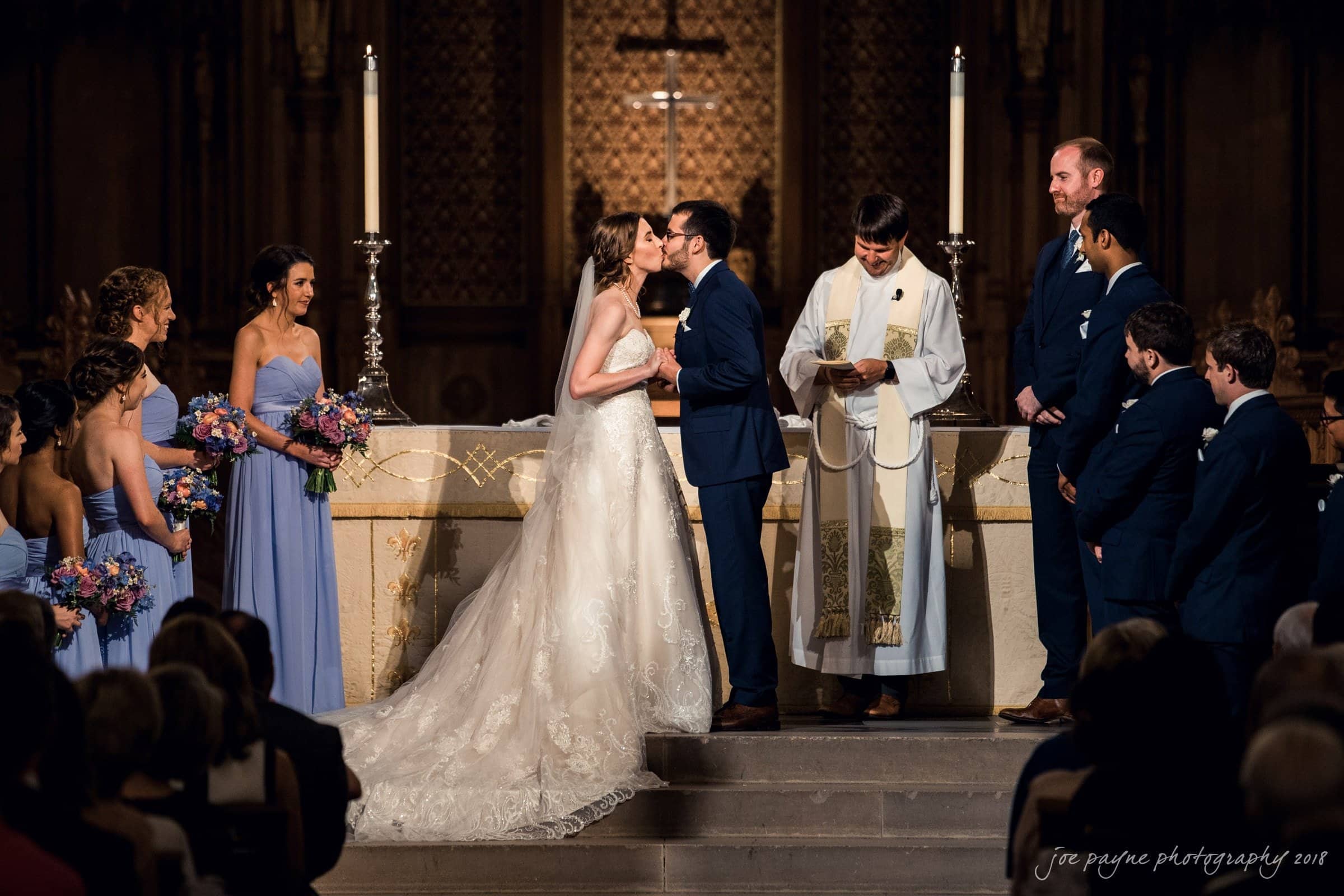 A bride and groom kiss at the altar during their wedding ceremony in a church. Bridesmaids in lavender dresses and groomsmen in dark suits stand by. A priest officiates, holding a book. Flowers decorate the scene.