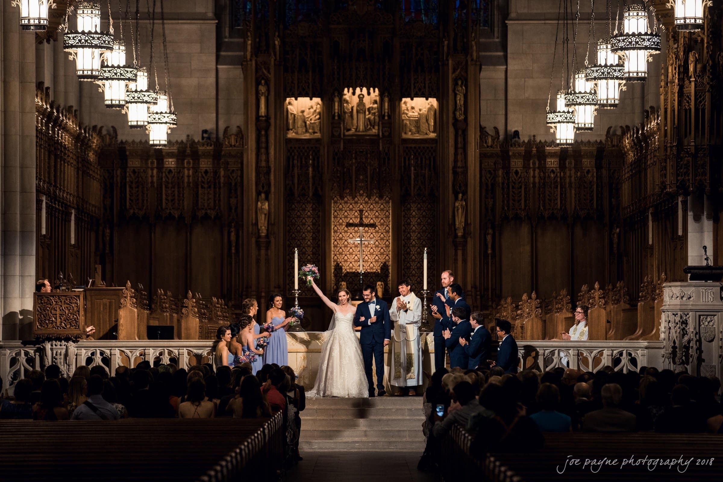 A bride and groom stand at the altar in a grand cathedral during their wedding ceremony. Bridesmaids and groomsmen flank them on either side, all dressed in formal attire. Guests are seated in the pews, witnessing the event.