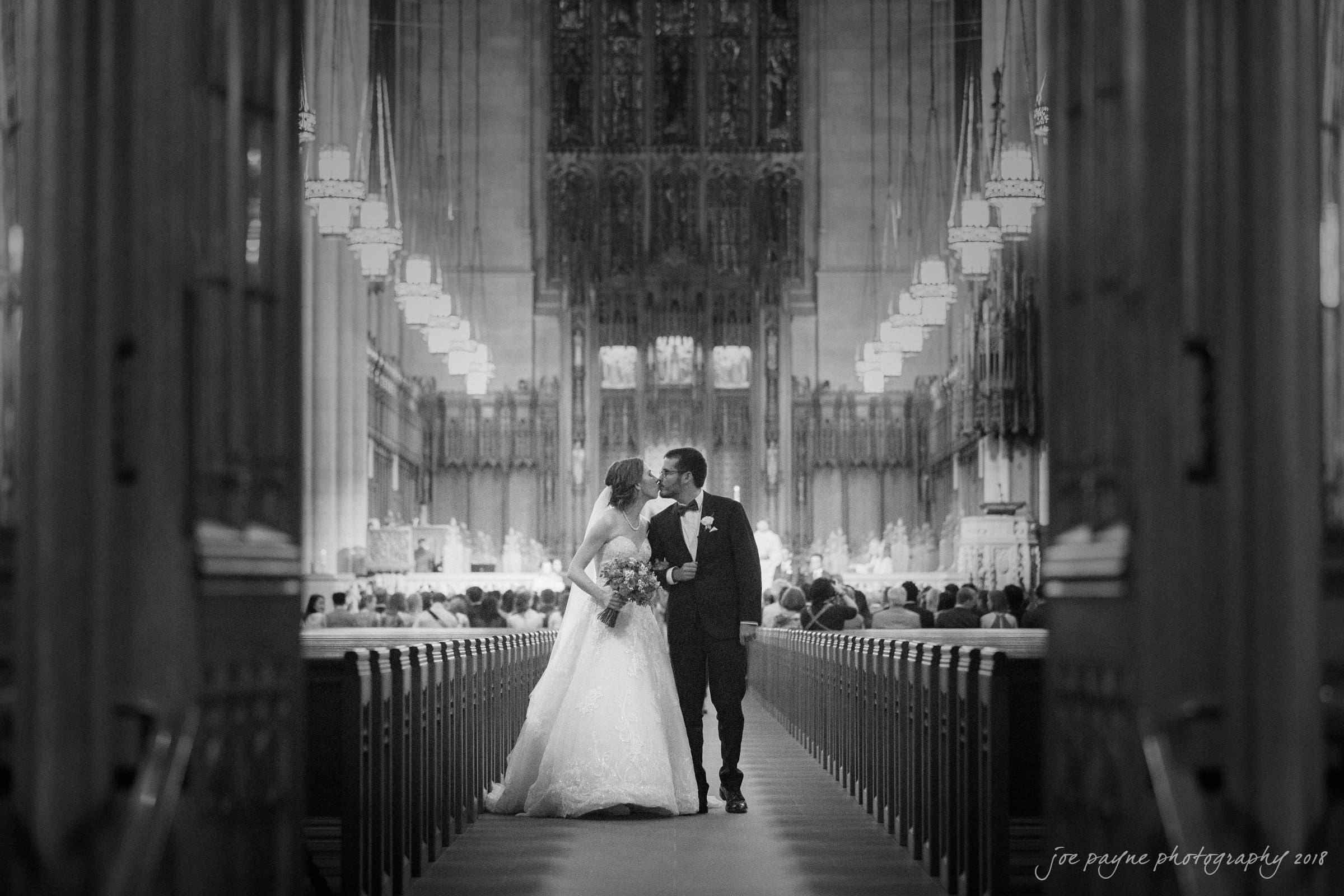 A bride and groom walk arm in arm down the aisle of a grand cathedral, holding hands and smiling at each other. The cathedrals ornate architectural details are visible in the background. The setting is elegant and atmospheric.