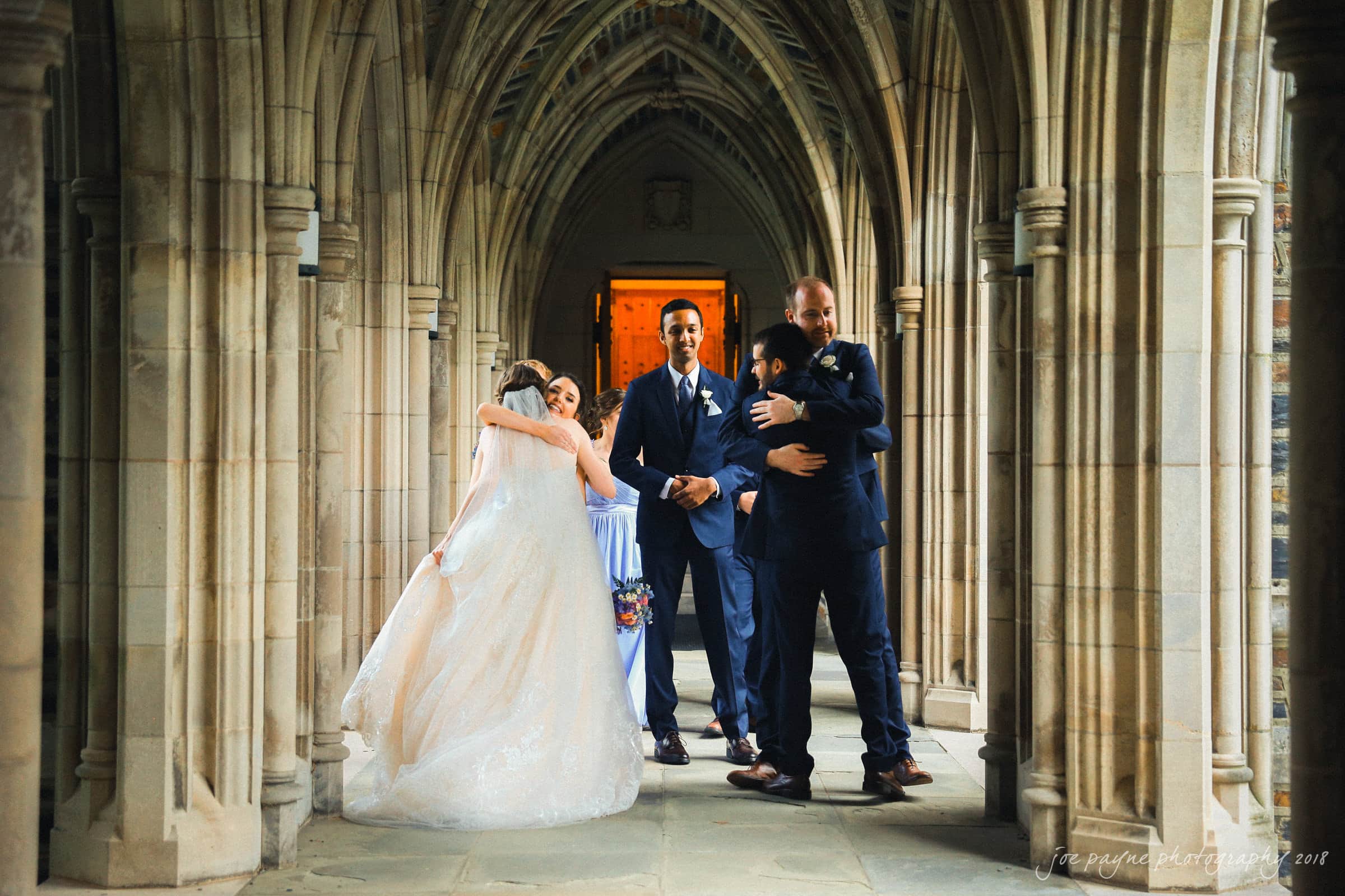 A bride and groom hug guests in a stone hallway with arched ceilings. The bride wears a white gown, and the groom is in a dark suit. Other guests in suits and dresses are embracing in the background.