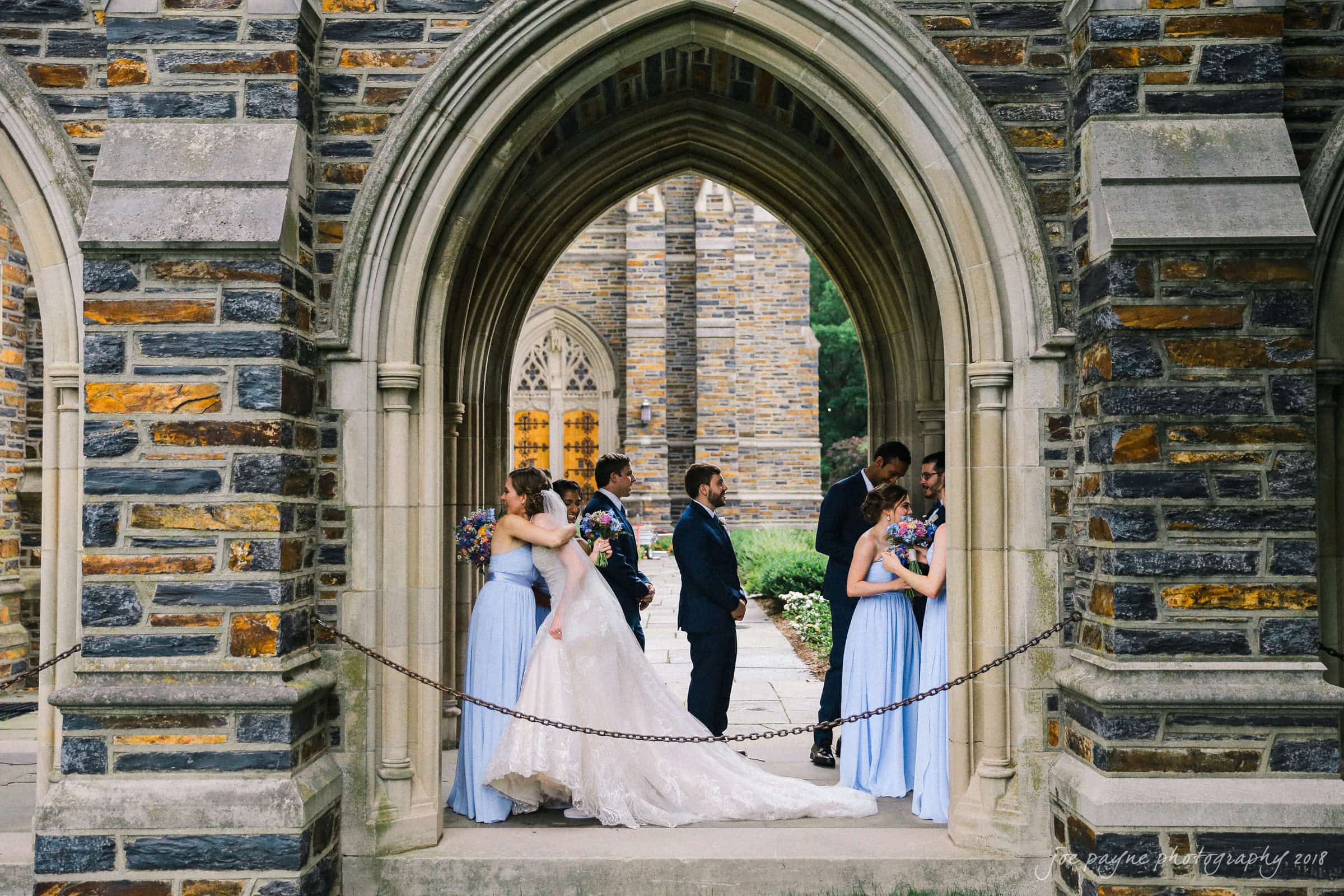 A bride and groom share a kiss under an archway, surrounded by bridesmaids in light blue dresses and groomsmen in dark suits. The stone architecture provides a classic backdrop.