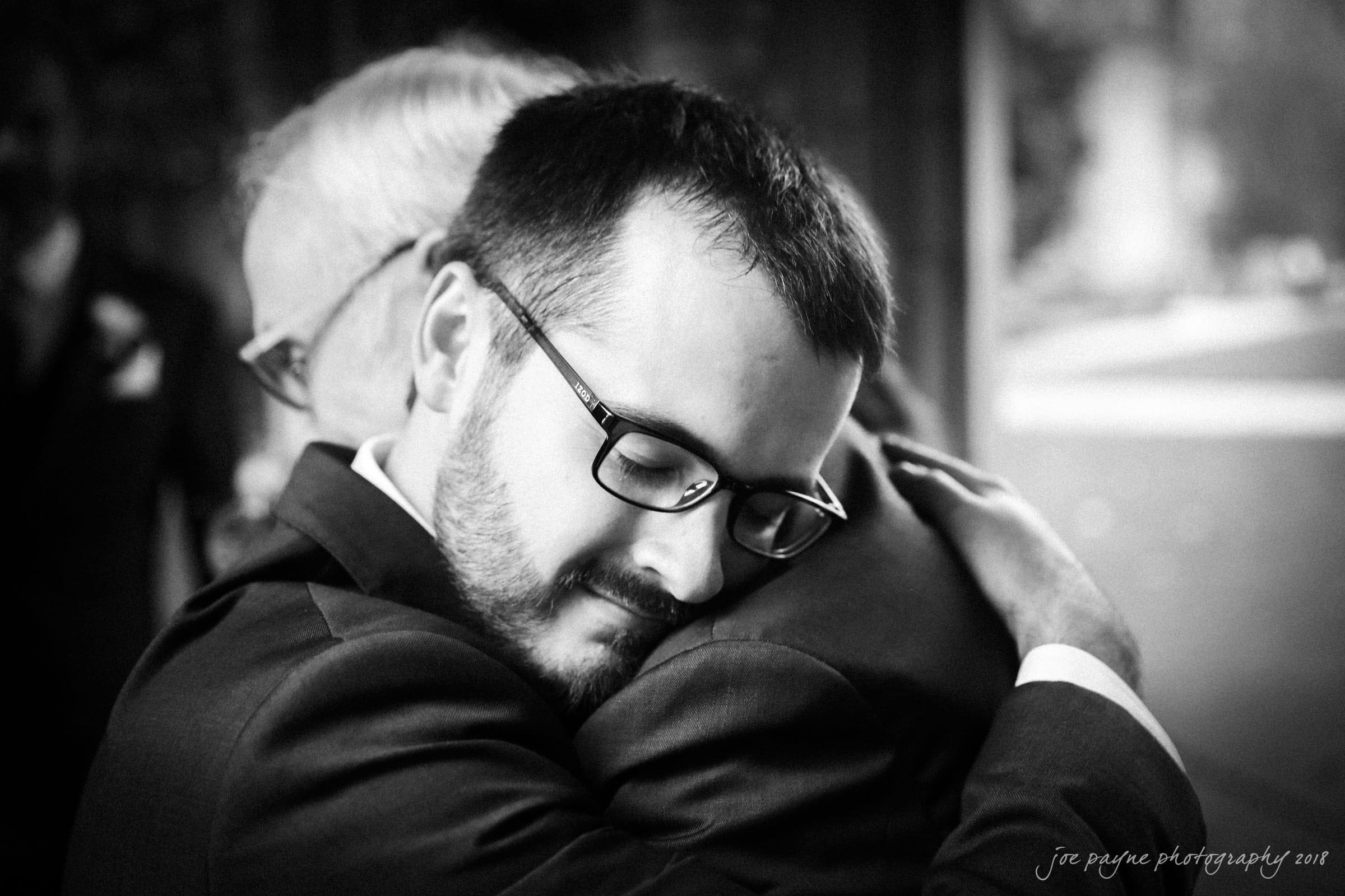 A black-and-white image of two men embracing. One man, wearing glasses and a suit, has his eyes closed, expressing emotion. The other mans face is partially visible as he hugs back. The background is slightly blurred.
