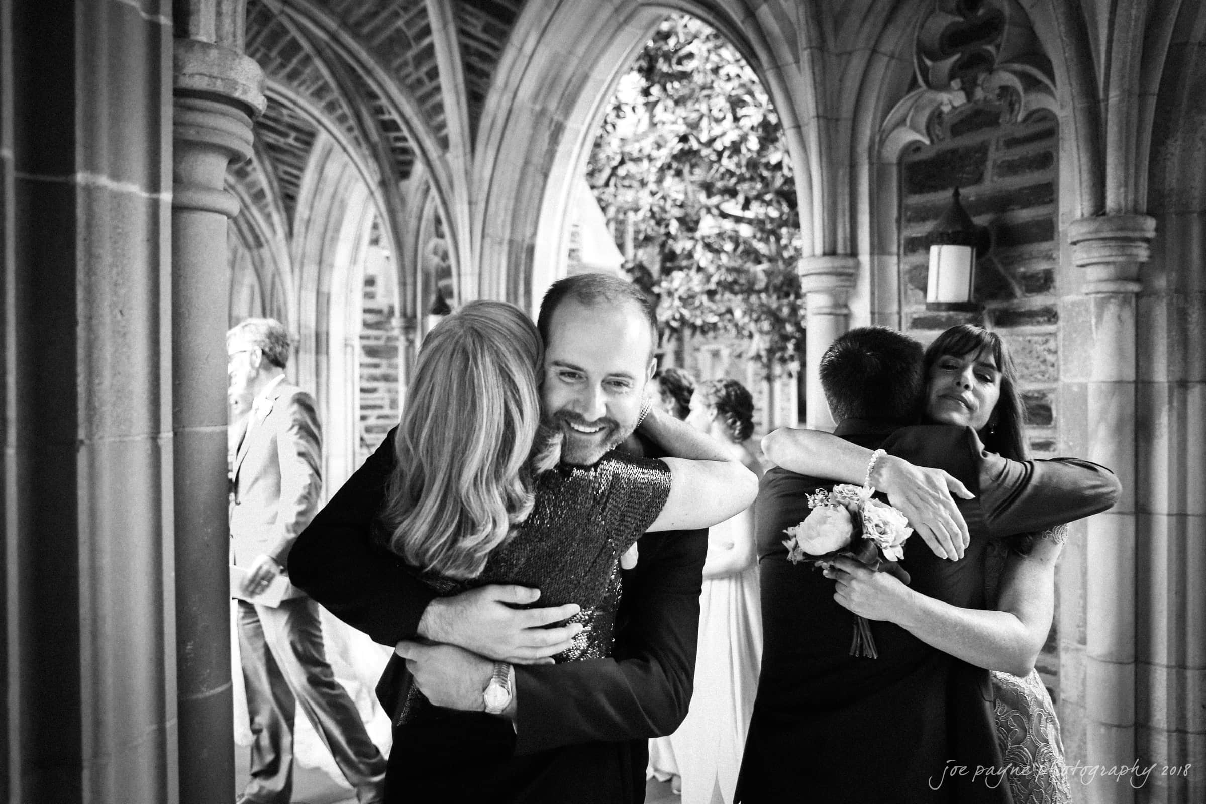 A black and white photo of two couples embracing in a corridor with stone arches. One couple, a man in a suit and a woman in a dress, are hugging and smiling. Another couple hugs beside them, one holding a bouquet. Others converse in the background.