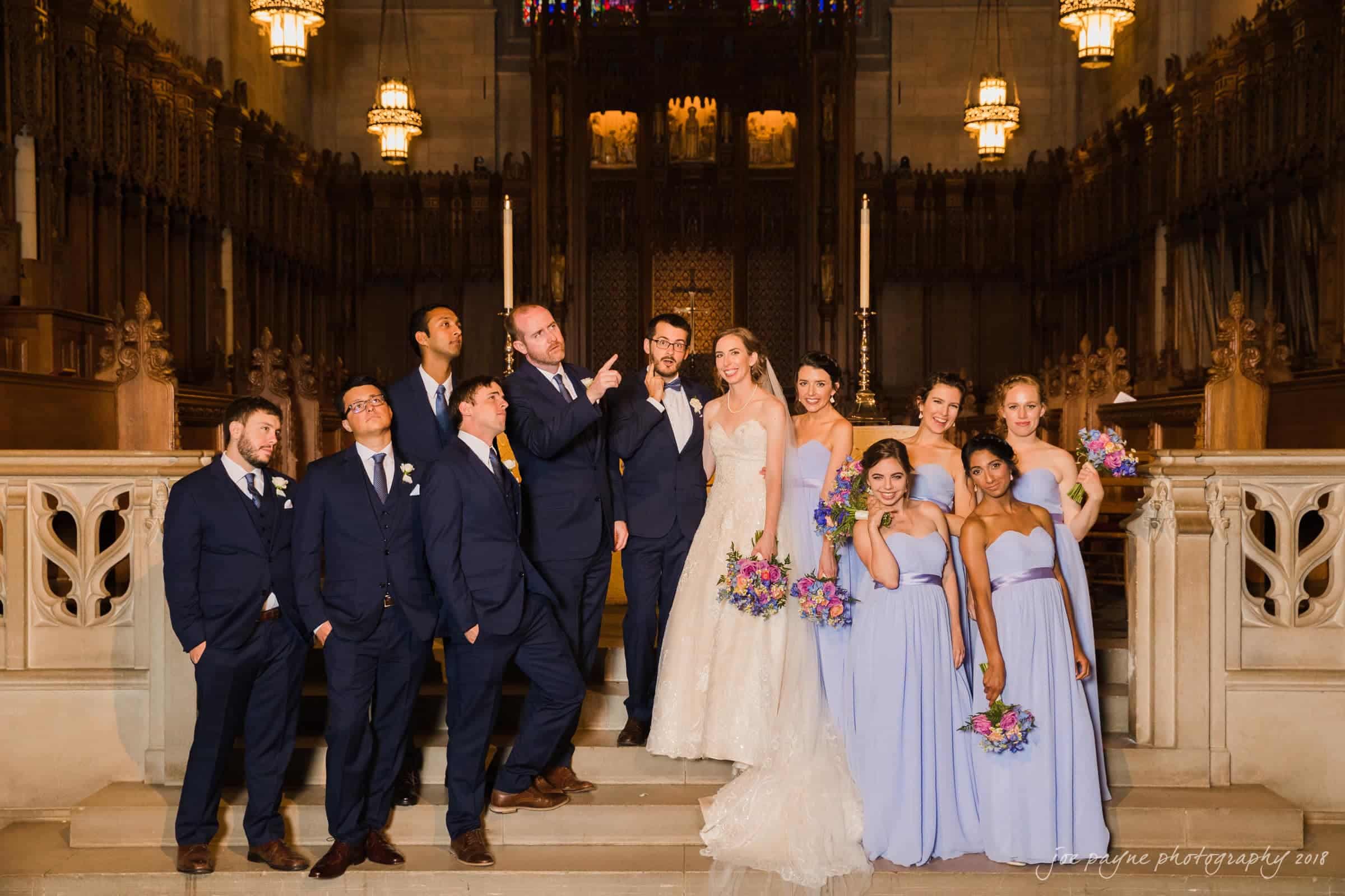 A wedding party poses on a staircase inside a church. The bride in a white gown and the groom in a dark suit stand at the center, surrounded by bridesmaids in lavender dresses and groomsmen in dark suits. Stained glass and a wooden interior are visible.