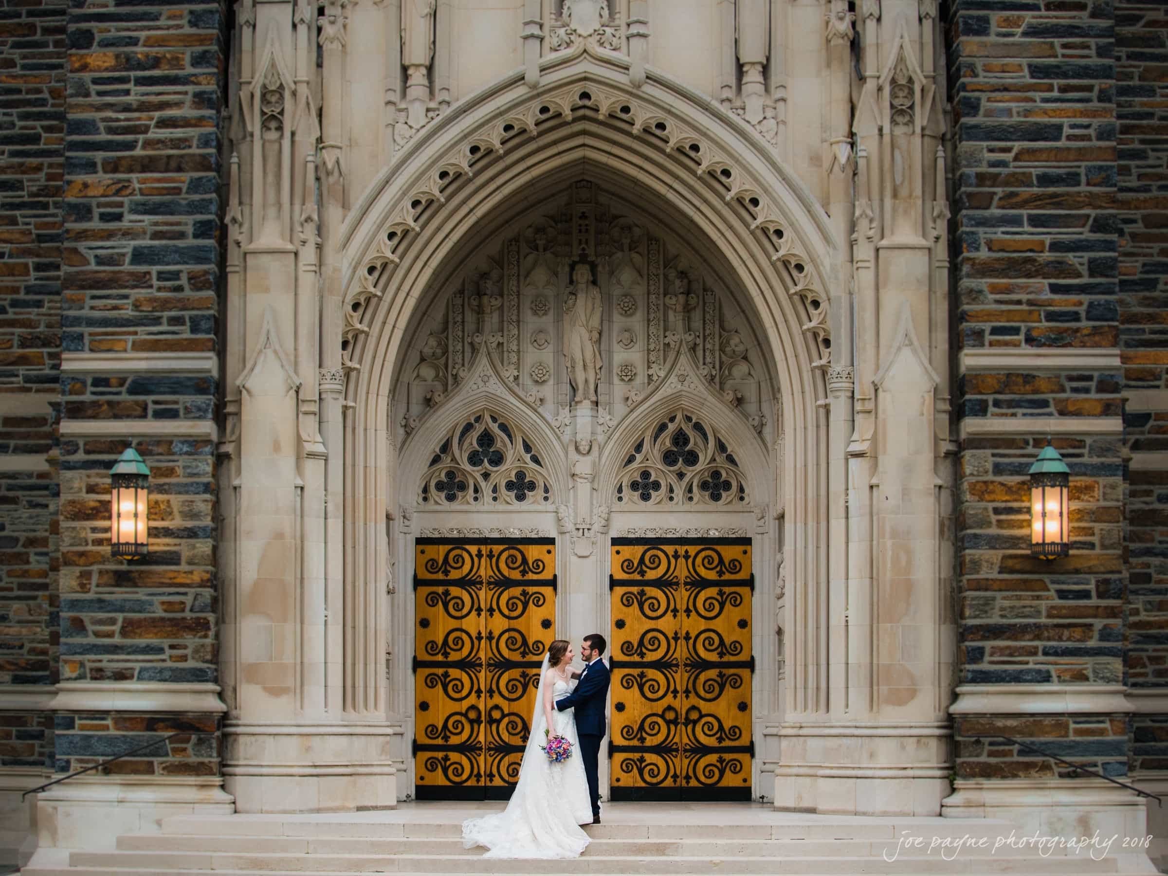 A bride and groom stand in front of a grand, ornately carved church entrance. The bride holds a bouquet, and they gaze at each other, framed by the cathedrals decorative stone archway and large, ornate doors.
