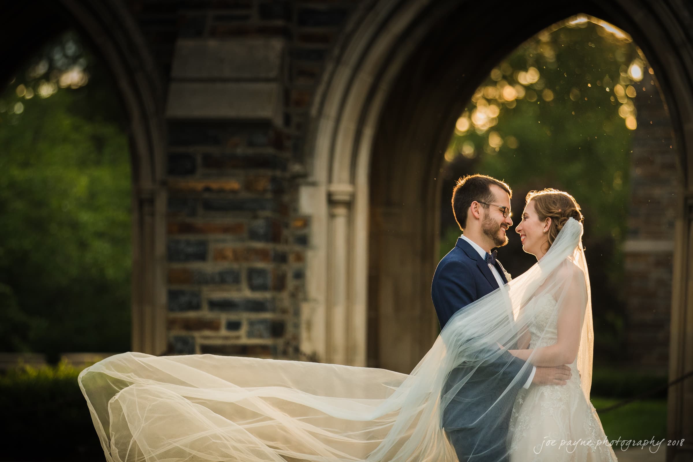 A bride and groom stand in front of an arched stone structure, smiling at each other. The brides veil flows elegantly in the breeze. Sunlight filters through the trees in the background, creating a warm, romantic atmosphere.