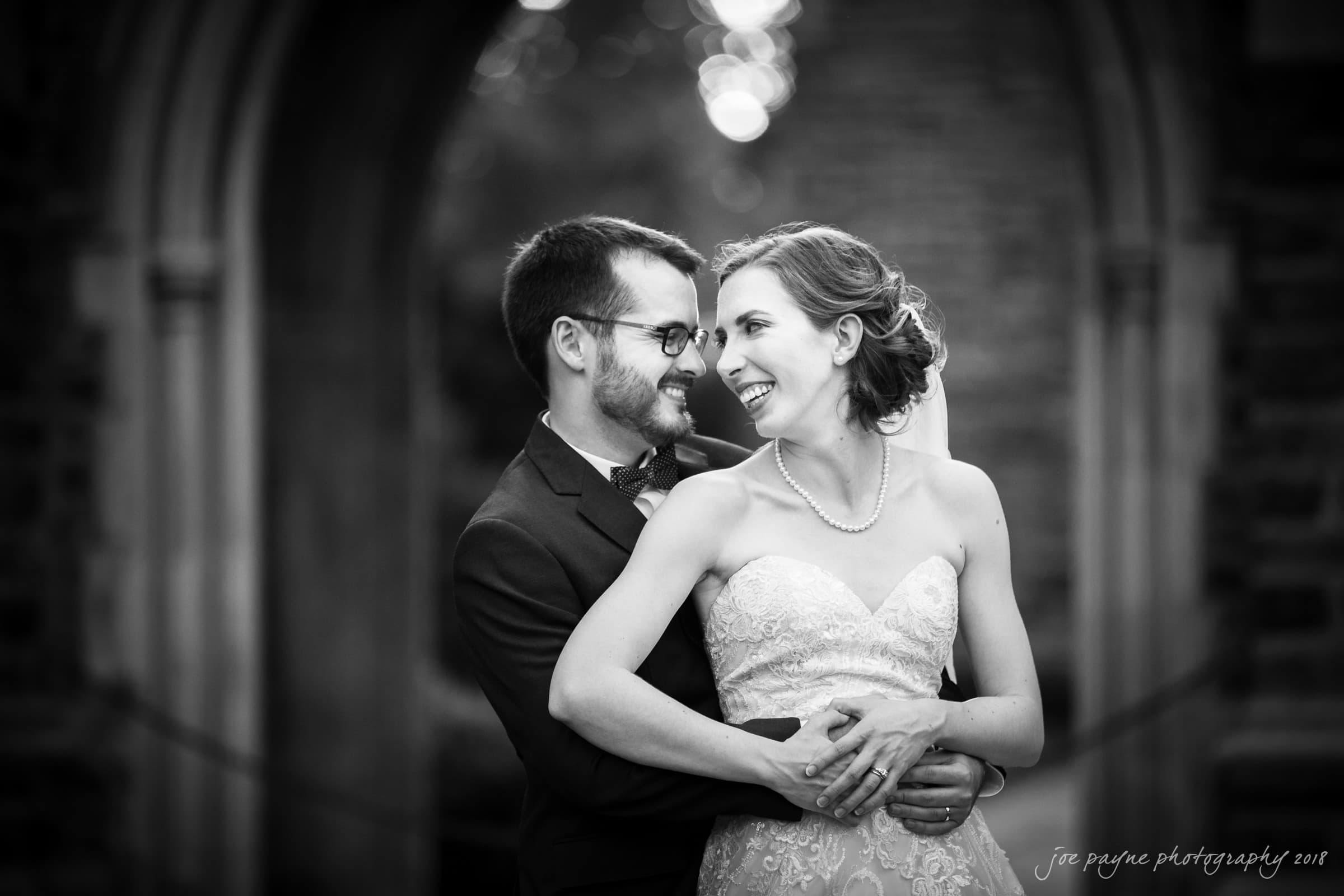 A black and white photo of a couple in formal attire. The man in a suit embraces the woman in a strapless gown. They are smiling at each other under an archway with blurred trees in the background.