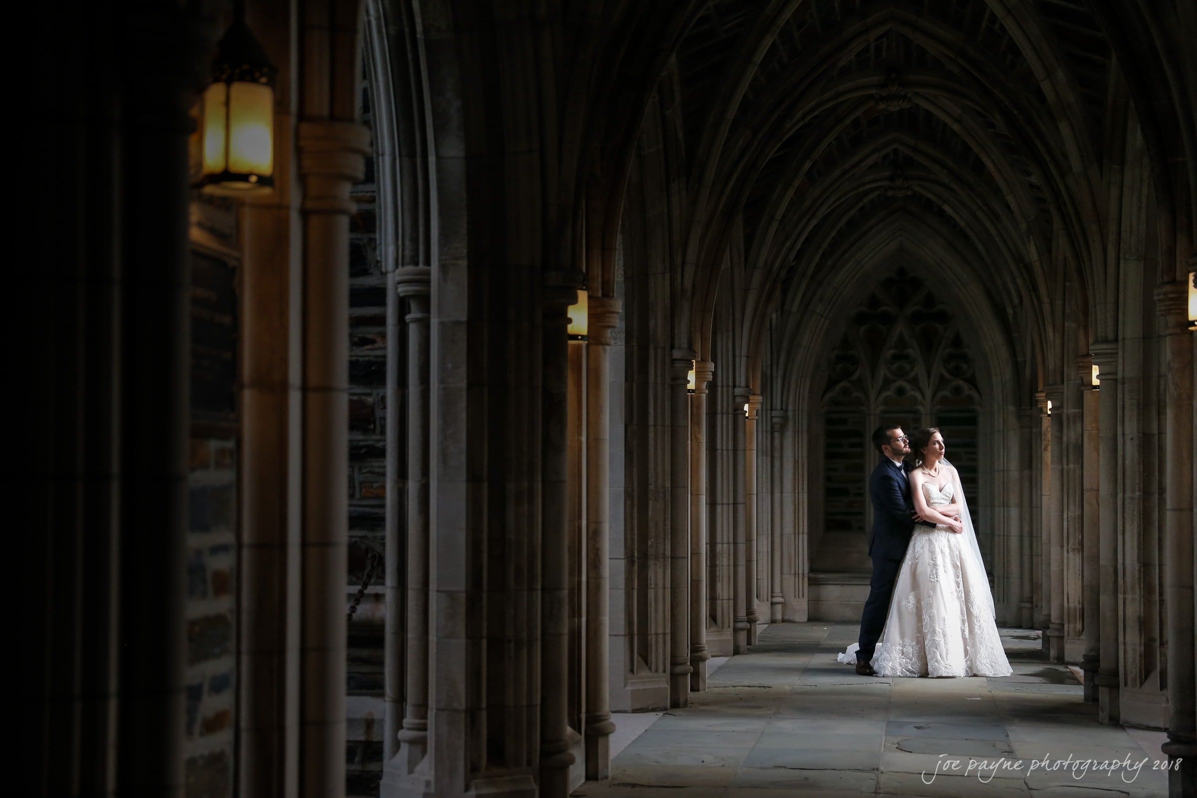 A couple in formal attire stands closely together in a dimly lit, arched stone corridor. Soft light from wall lanterns highlights the intricate design of the vaulted ceiling and the brides gown, adding a romantic ambiance to the scene.