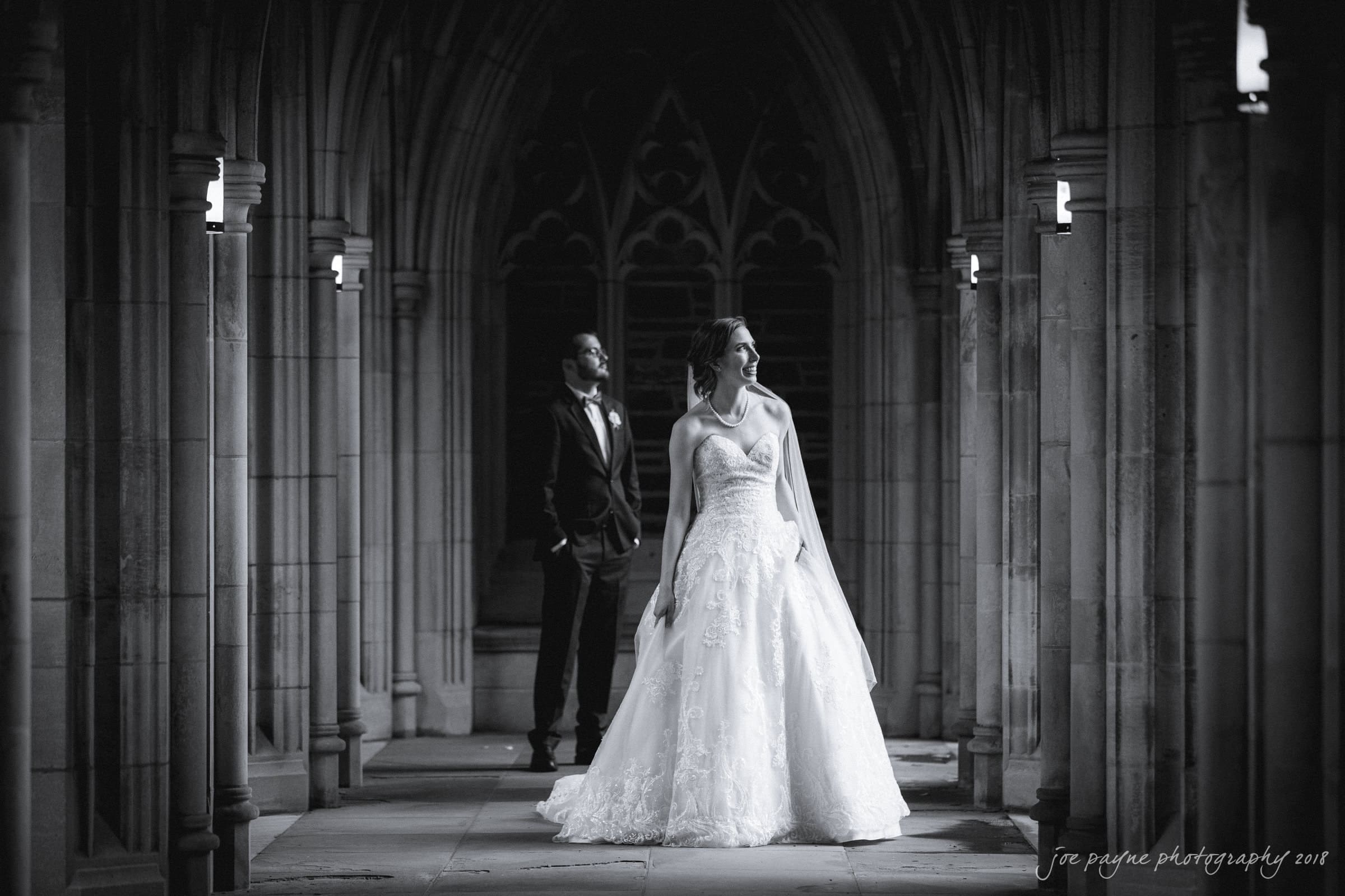 A bride in an elegant gown poses in a stone hallway with archways. A groom in a tuxedo stands behind her, looking away. The scene is lit by small wall lamps, creating a romantic and timeless atmosphere.