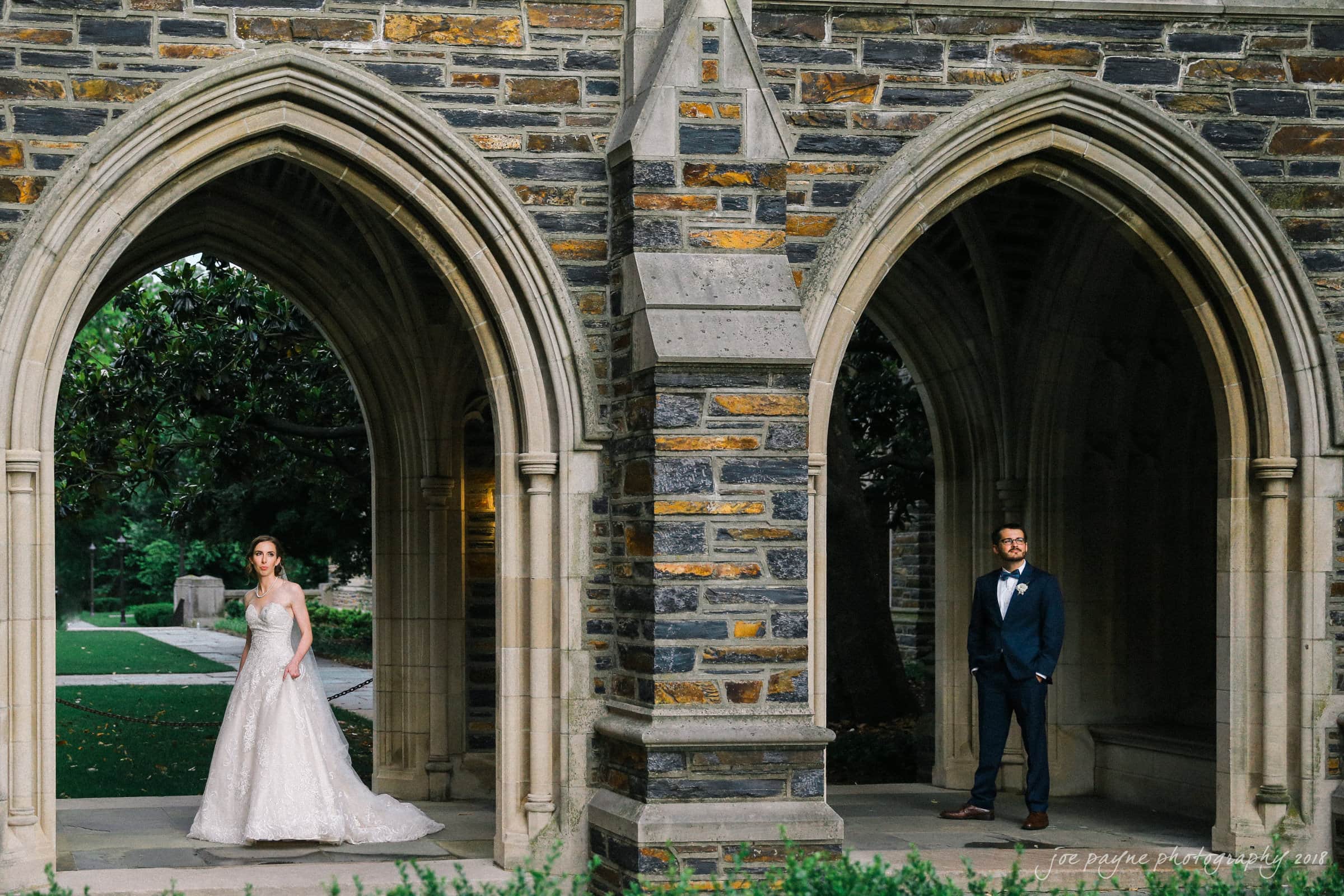 A bride in a white gown and a groom in a suit stand in separate stone archways of a historic building, facing each other. The background features greenery and intricate stonework.