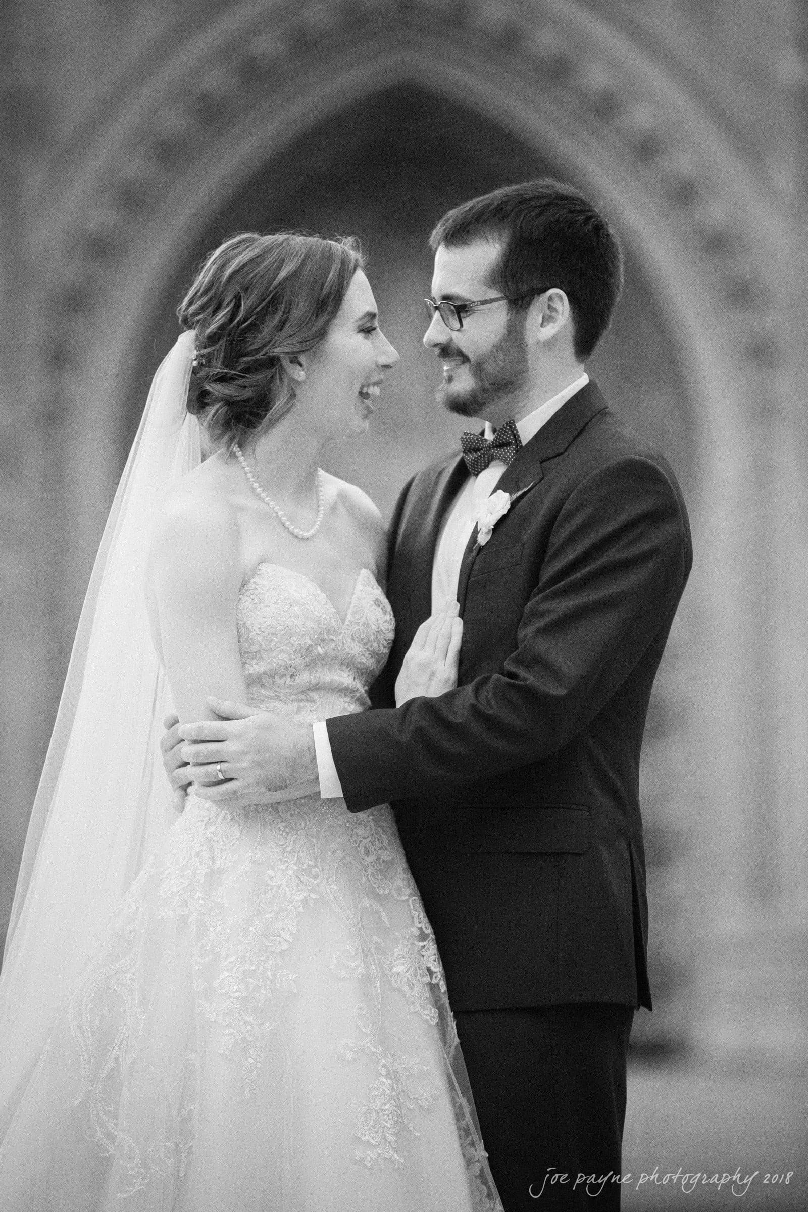 A black and white photo of a bride and groom standing together. The bride wears a strapless lace wedding dress and a veil. The groom is in a suit with a bow tie. They are looking at each other and smiling, with an ornate archway in the background.