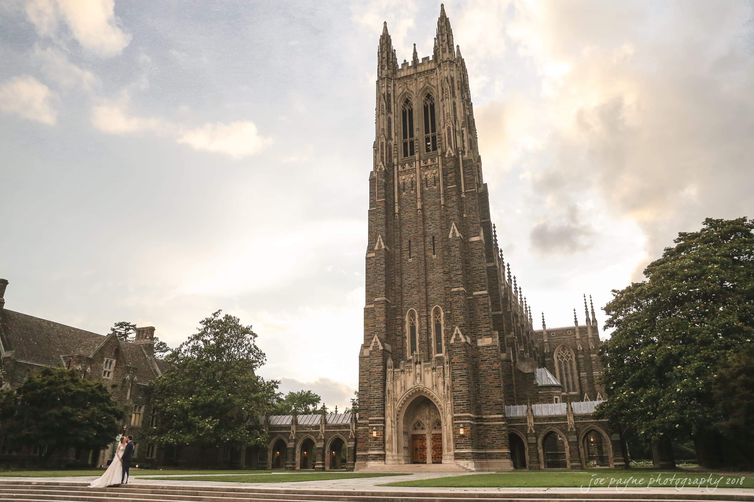 A couple stands in front of a large, Gothic-style cathedral with a tall tower, surrounded by grass and trees under a partly cloudy sky at sunset.