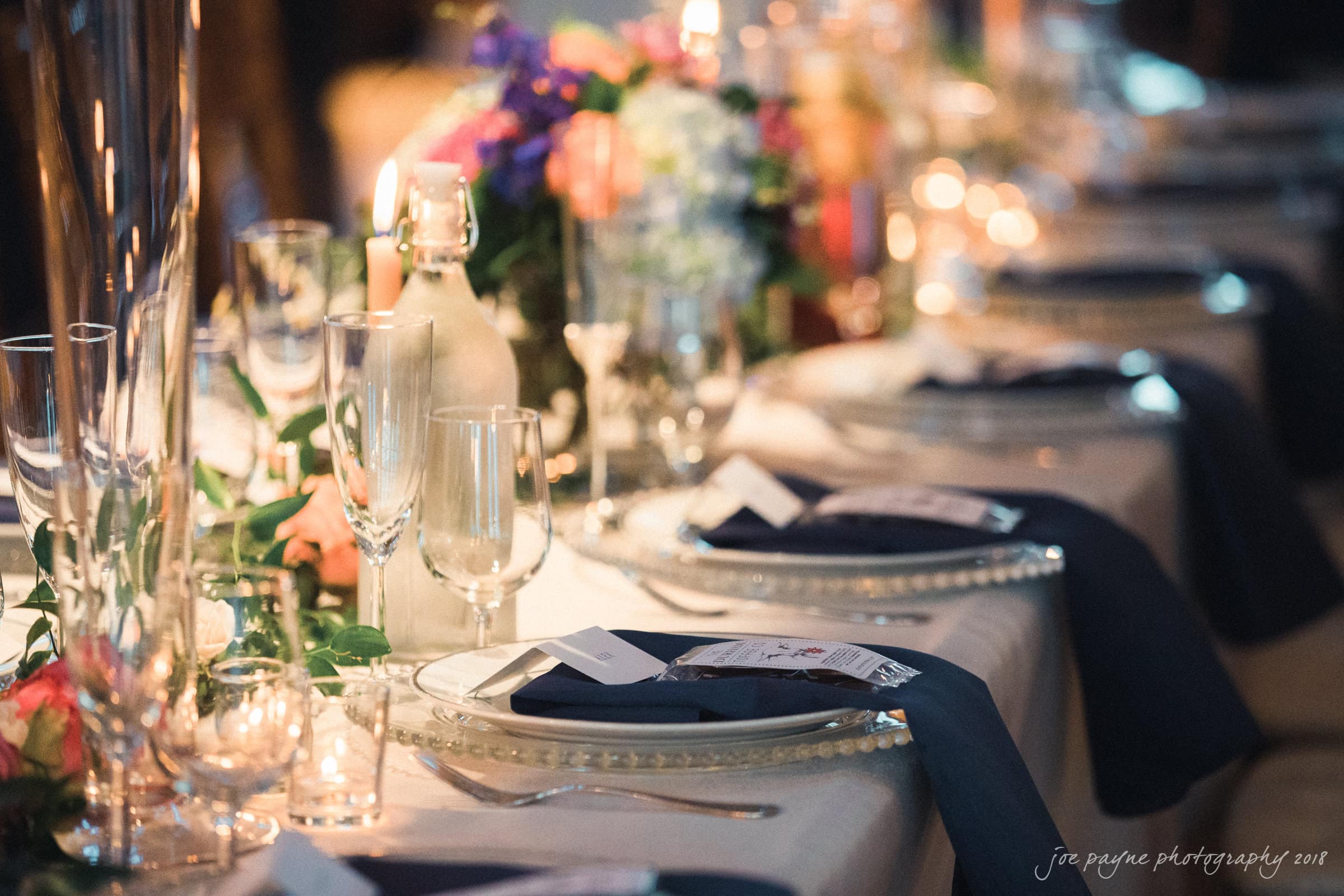 A beautifully set dinner table with elegant glassware, plates, and a dark napkin at each setting. Floral centerpieces and candles add warmth and color, creating an inviting atmosphere.
