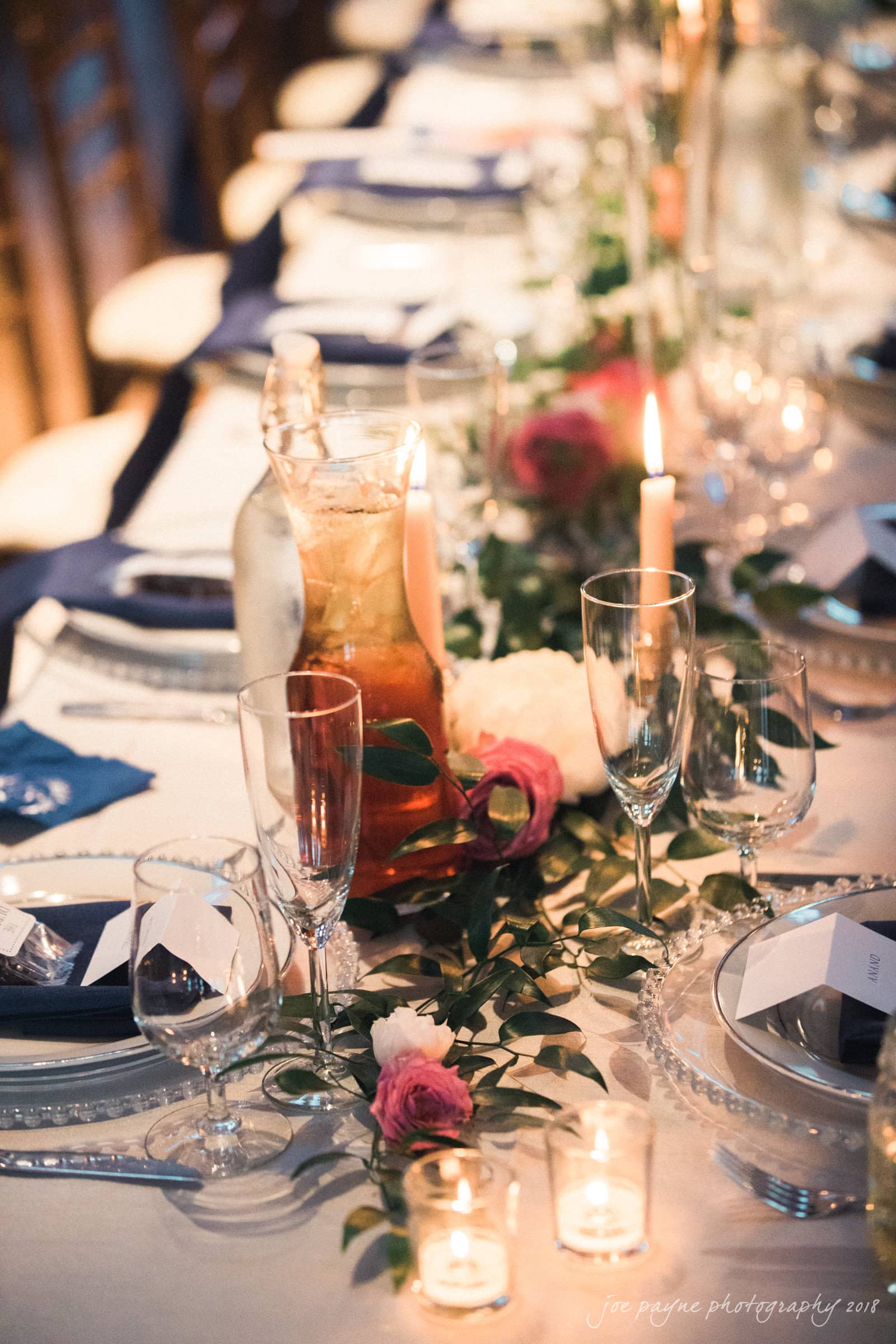 A beautifully set table for a formal event, featuring elegant glassware, plates, and lit candles. A floral centerpiece with greenery and pink roses runs along the center. A tall glass pitcher with a beverage is also present.