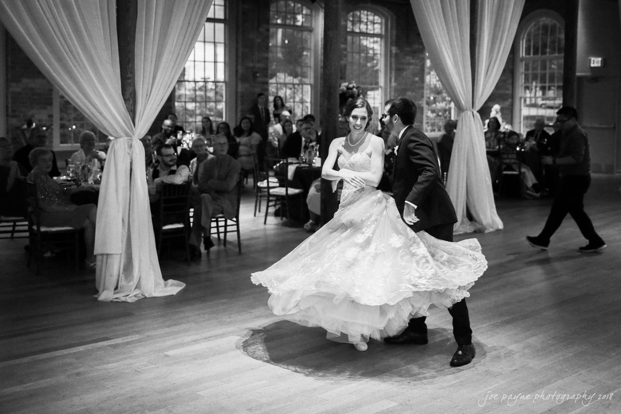 A bride and groom dance elegantly on a wooden floor in a large hall with draped curtains and spectators seated around. The brides dress swirls as they move. Guests watch from the background through large windows.