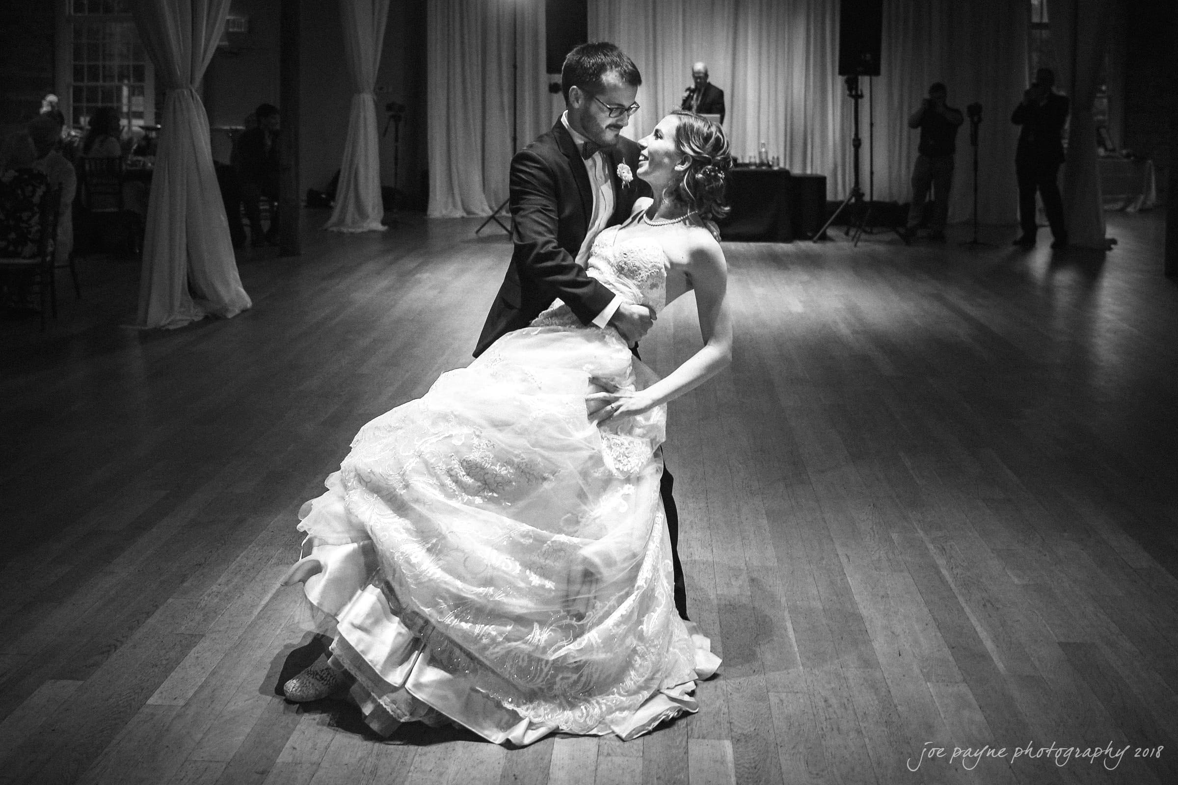 A couple performs a dramatic dance in an elegant setting. The man supports the woman in a dip, her flowing gown spread gracefully on the floor. Soft lighting and draped curtains enhance the romantic ambiance.