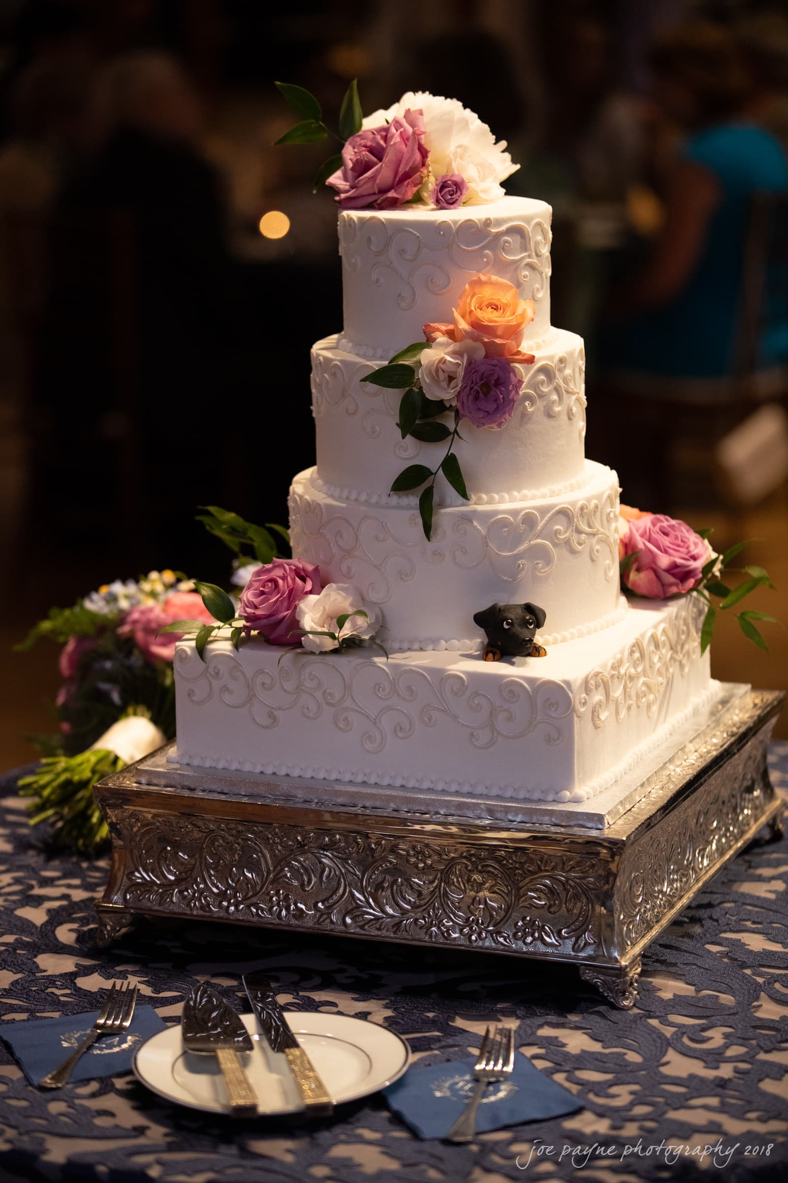 A four-tiered white wedding cake decorated with pink and purple roses, and greenery. It sits on an ornate silver stand on a blue lace tablecloth. A small black dog figurine peeks out from the bottom tier. Plates and cutlery are in the foreground.