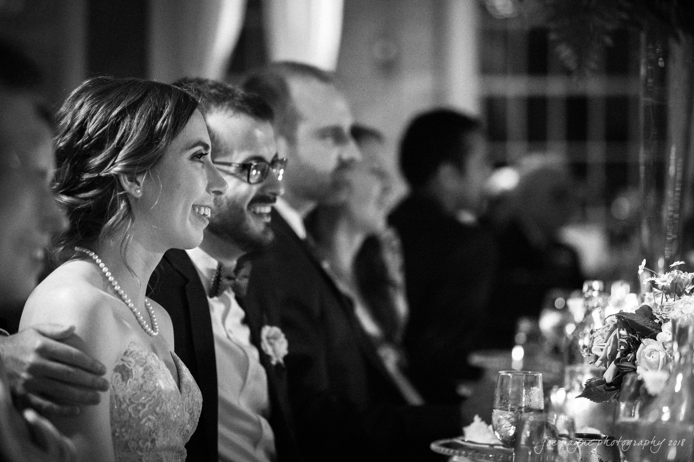 A black and white photo of a wedding reception. A bride and groom are seated at a table, smiling and looking towards an unseen speaker. The bride is wearing a lace dress and pearl necklace. Other guests are sitting nearby, with some flowers on the table.