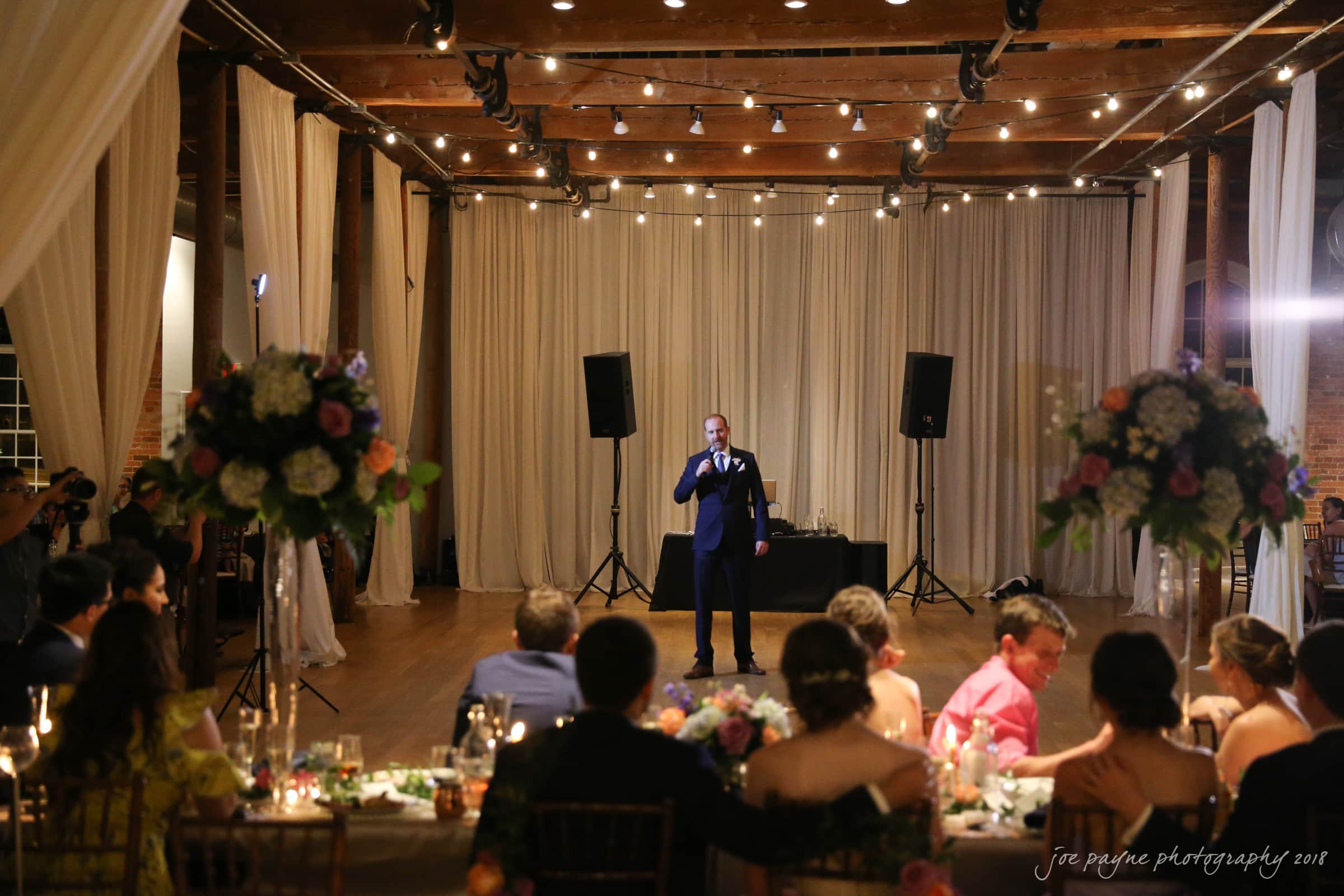 A man in a suit stands on a dance floor with a microphone, speaking to a seated audience at a wedding reception. The room is warmly lit with string lights and features elegant floral arrangements.