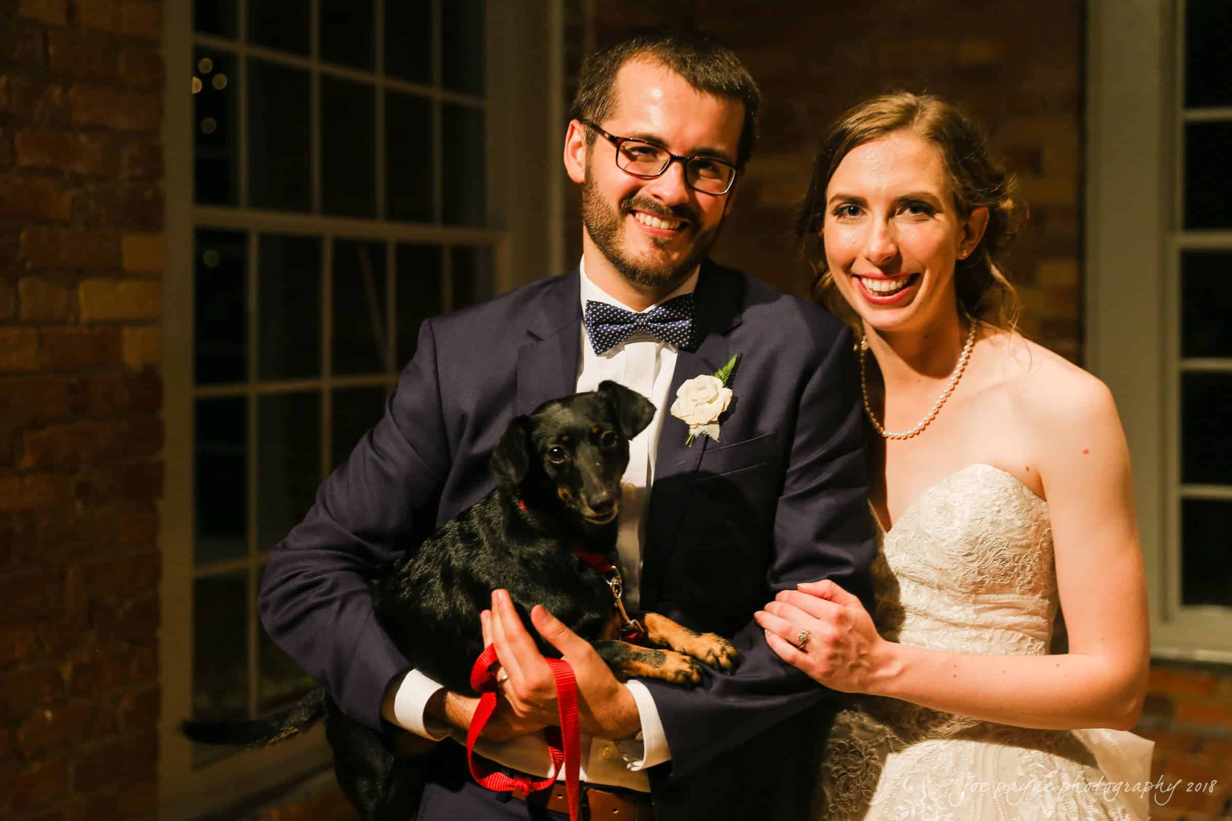 A smiling groom in a navy suit holds a small black dog with a red leash. A smiling bride in a white lace gown stands beside them. They pose by a window, and the setting appears warm and cozy.