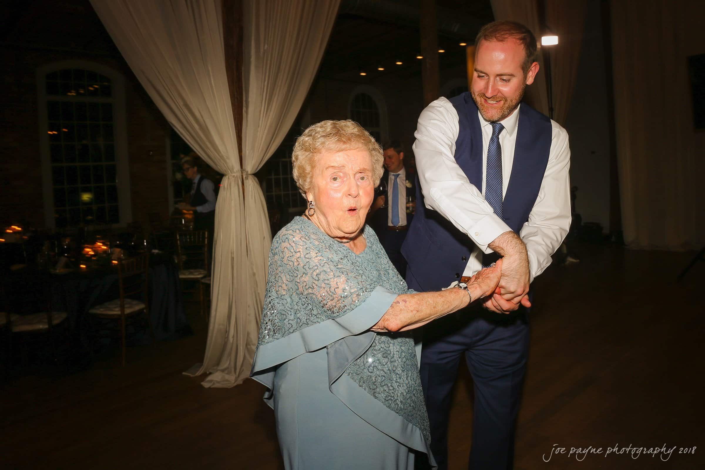An elderly woman with short hair dances joyfully with a bearded man in a suit and blue vest at an event. They are on a wooden dance floor with soft lighting and draped curtains in the background.