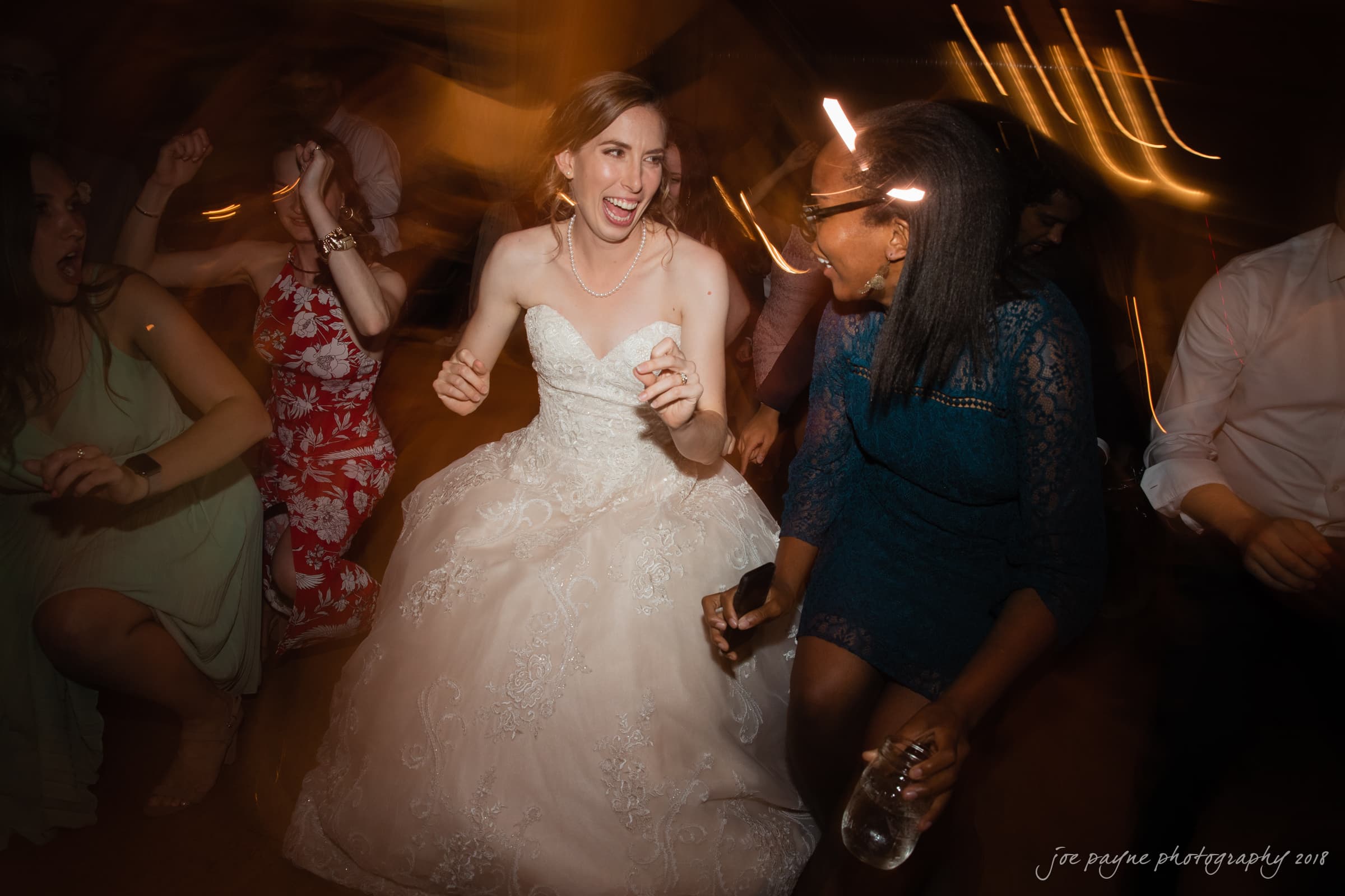 A bride in a white gown joyfully dances with guests at a wedding reception. The scene is lively, with people smiling and moving energetically around her. The lighting is warm and dim, creating a festive and celebratory atmosphere.