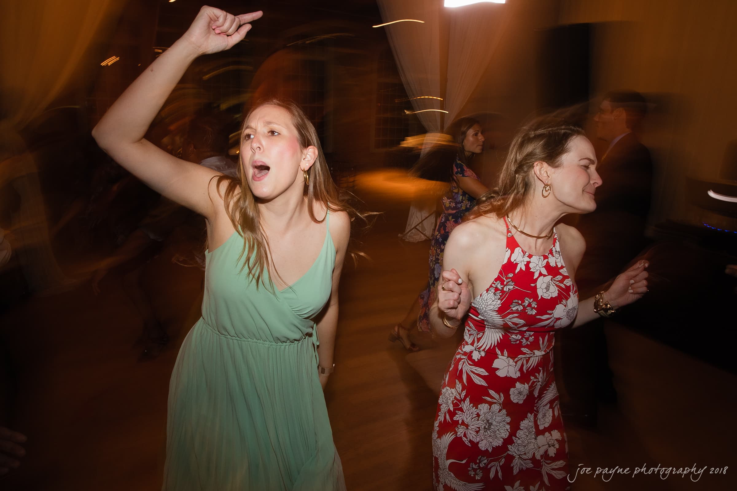 Two women are dancing energetically at a party. One is wearing a green dress, and the other a red floral dress. The scene is dimly lit, with motion blur conveying their movement and the lively atmosphere.