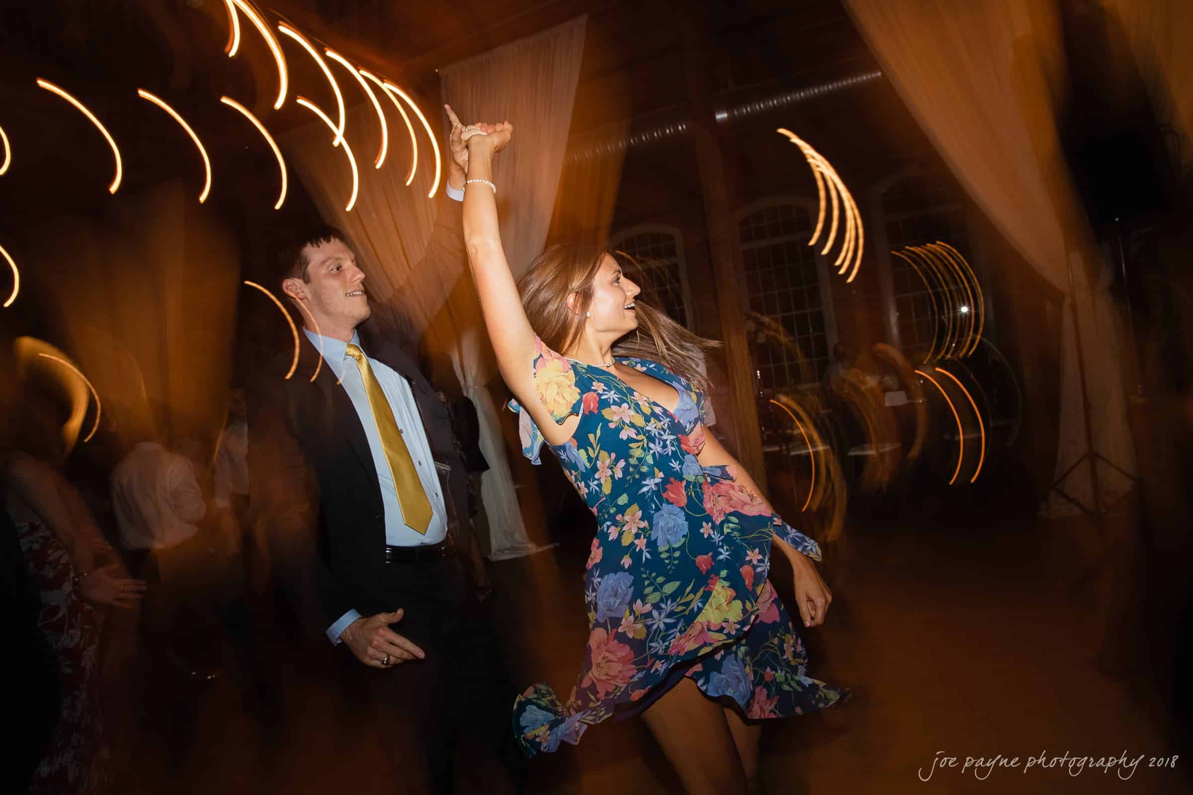 A man in a suit and a woman in a floral dress dance energetically, spinning under soft, swirling lights. The setting appears to be a dimly lit event or party, creating a lively and joyful atmosphere.