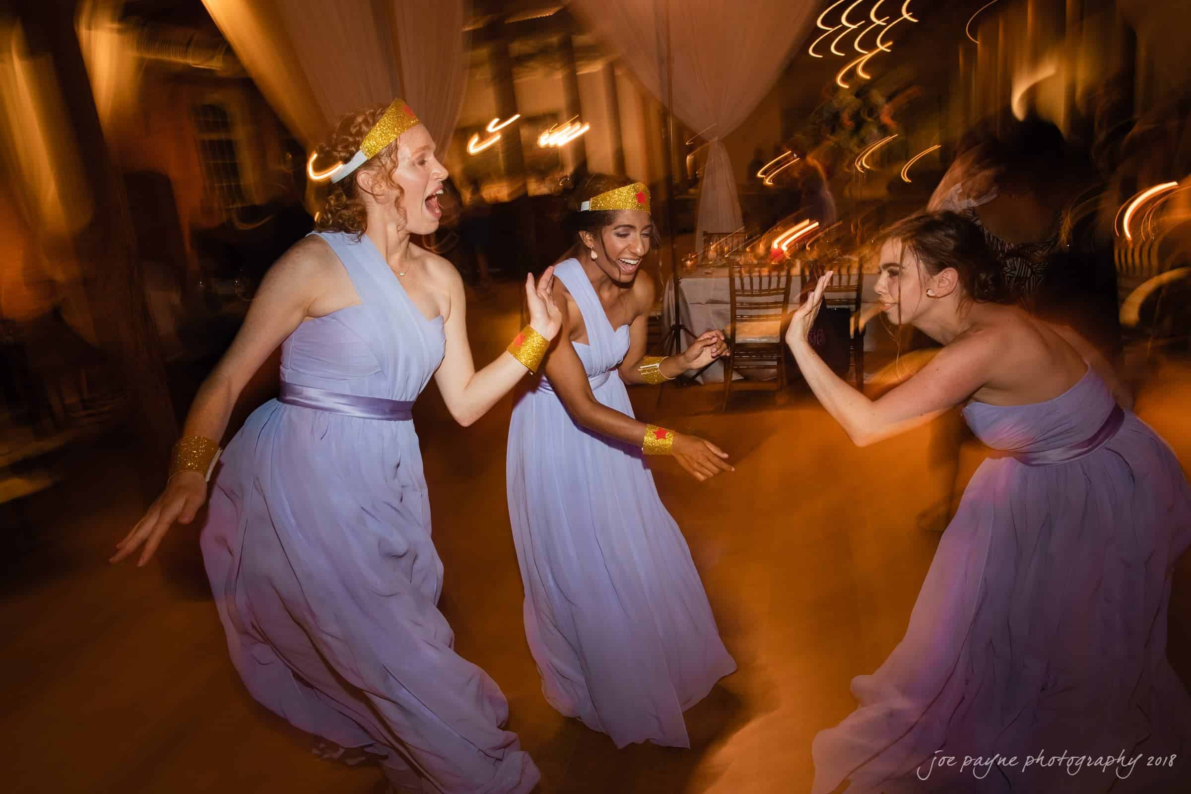 Three women in lavender dresses and gold accessories dance joyfully in a warmly lit room. The setting suggests a festive event. The image captures movement and energy with a blurry, dynamic effect.