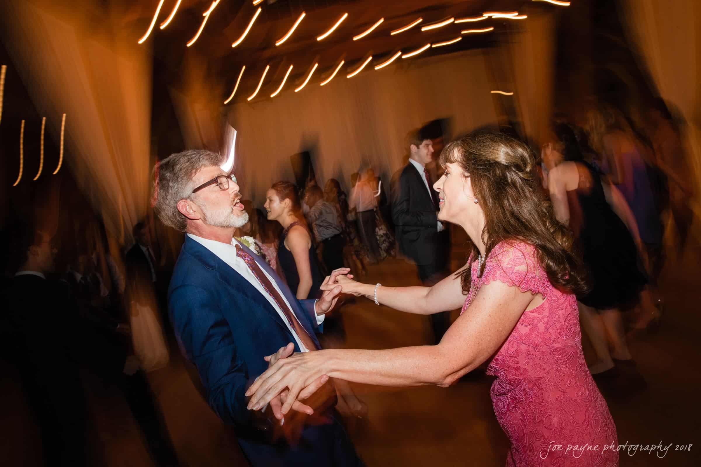 A man in a blue suit and a woman in a pink dress are joyfully dancing together at an event. The background is motion-blurred with warm lighting, creating a lively and festive atmosphere.