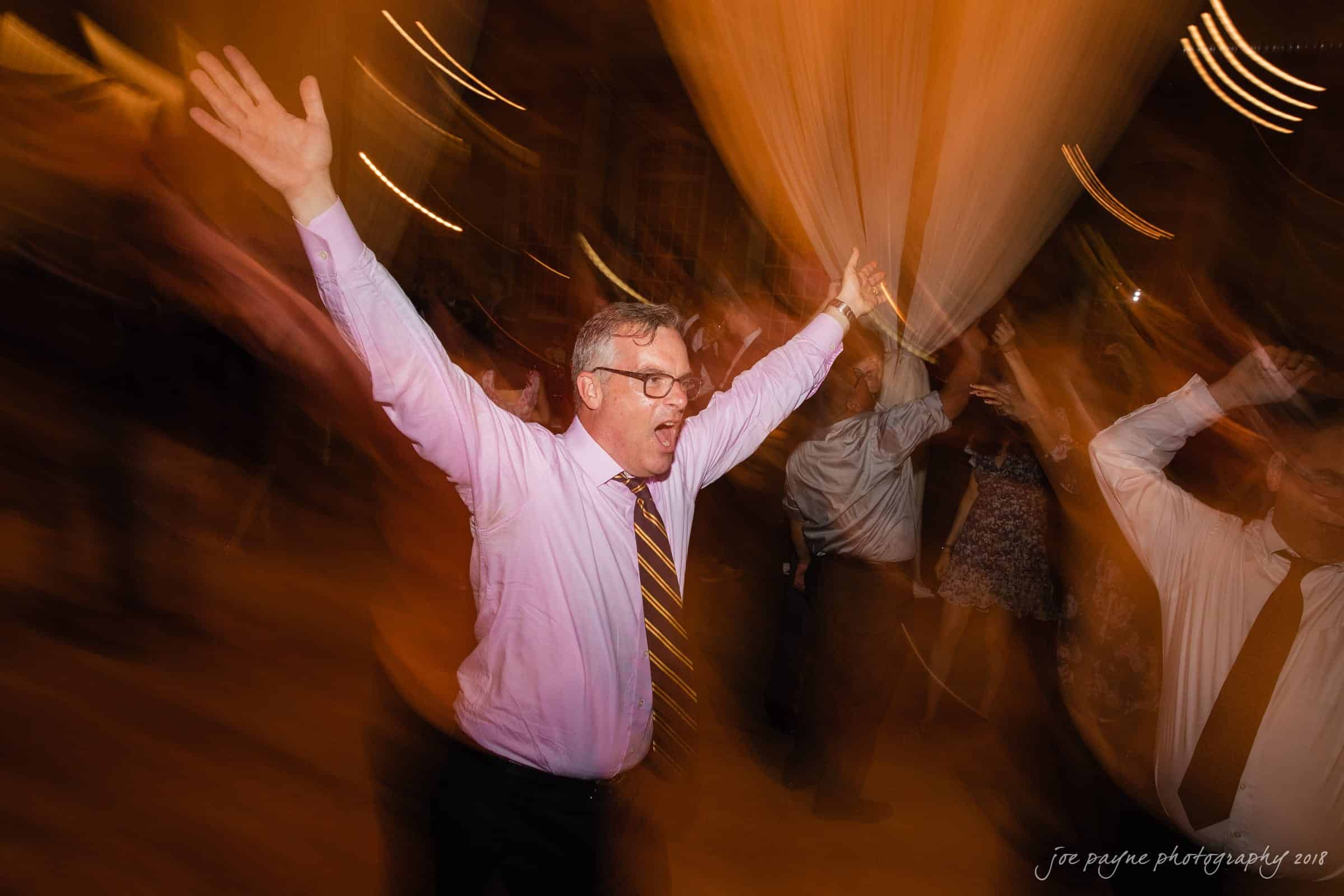 A man in a pink shirt and tie joyfully dances with his arms raised at an event. The background features blurred lights and other dancers, creating a dynamic and energetic atmosphere.