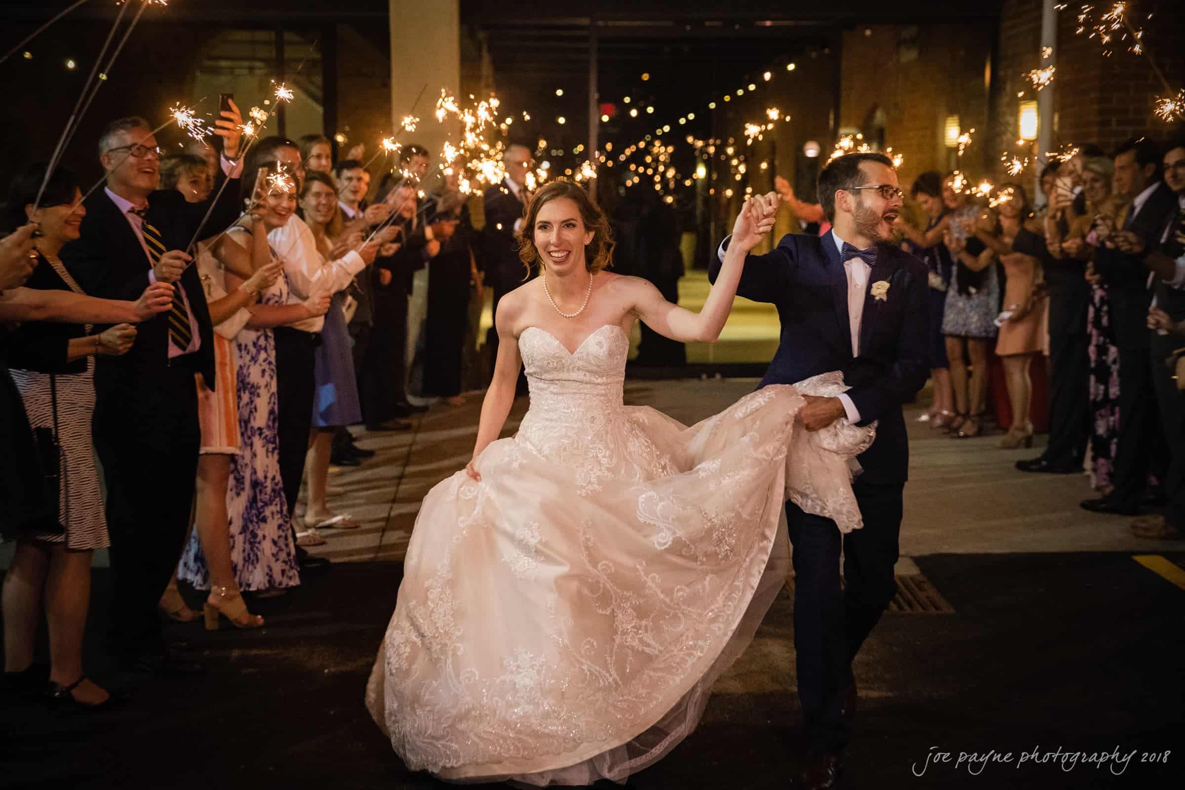 A bride and groom joyfully exit a building at night, holding hands under a canopy of sparklers held by cheering guests. The bride wears a white gown and the groom a dark suit. String lights are visible in the background.