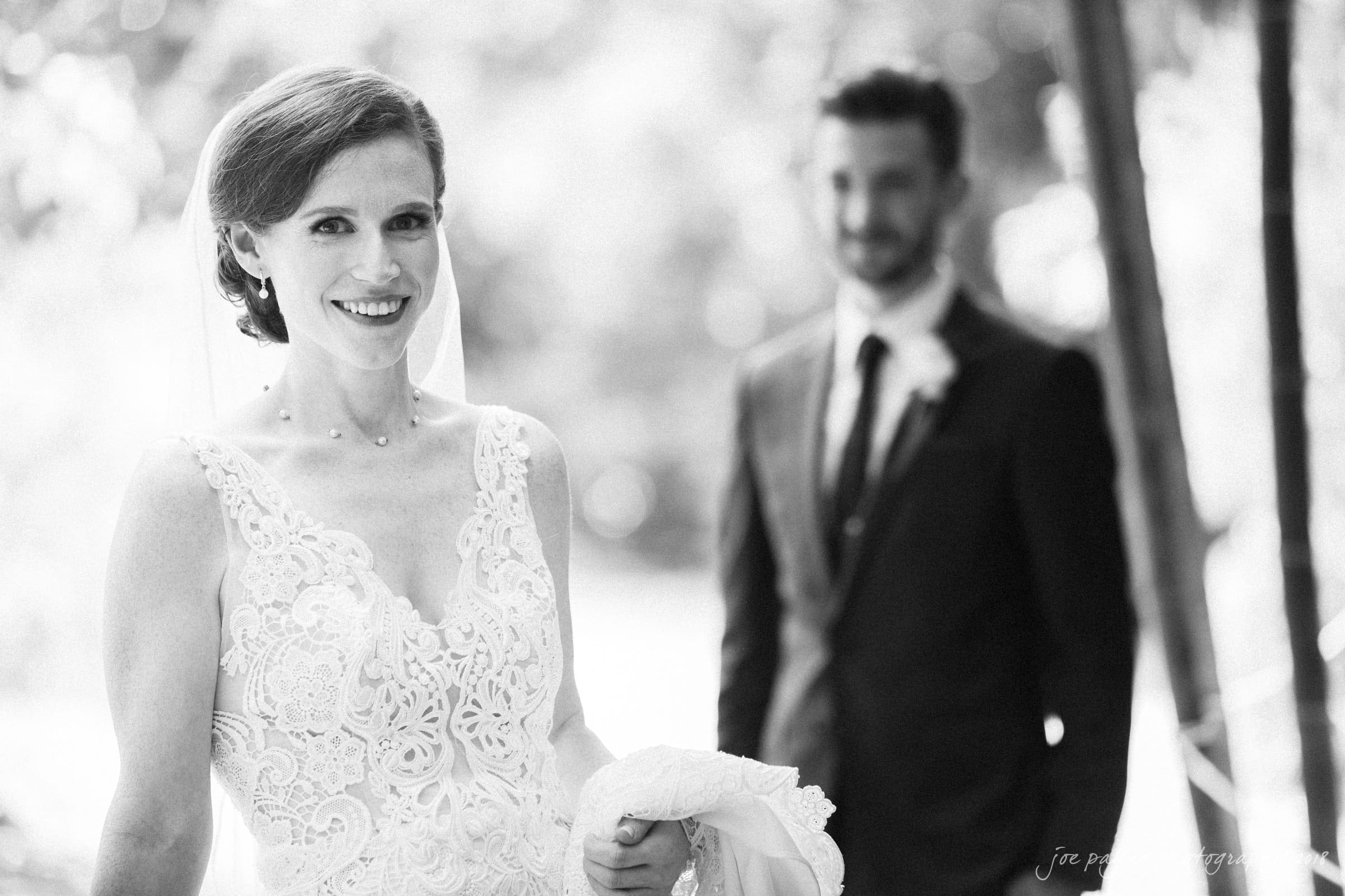 A black and white photo of a bride in a lace gown and veil, smiling at the camera. A groom in a suit stands slightly out of focus in the background. They are outdoors, surrounded by greenery.
